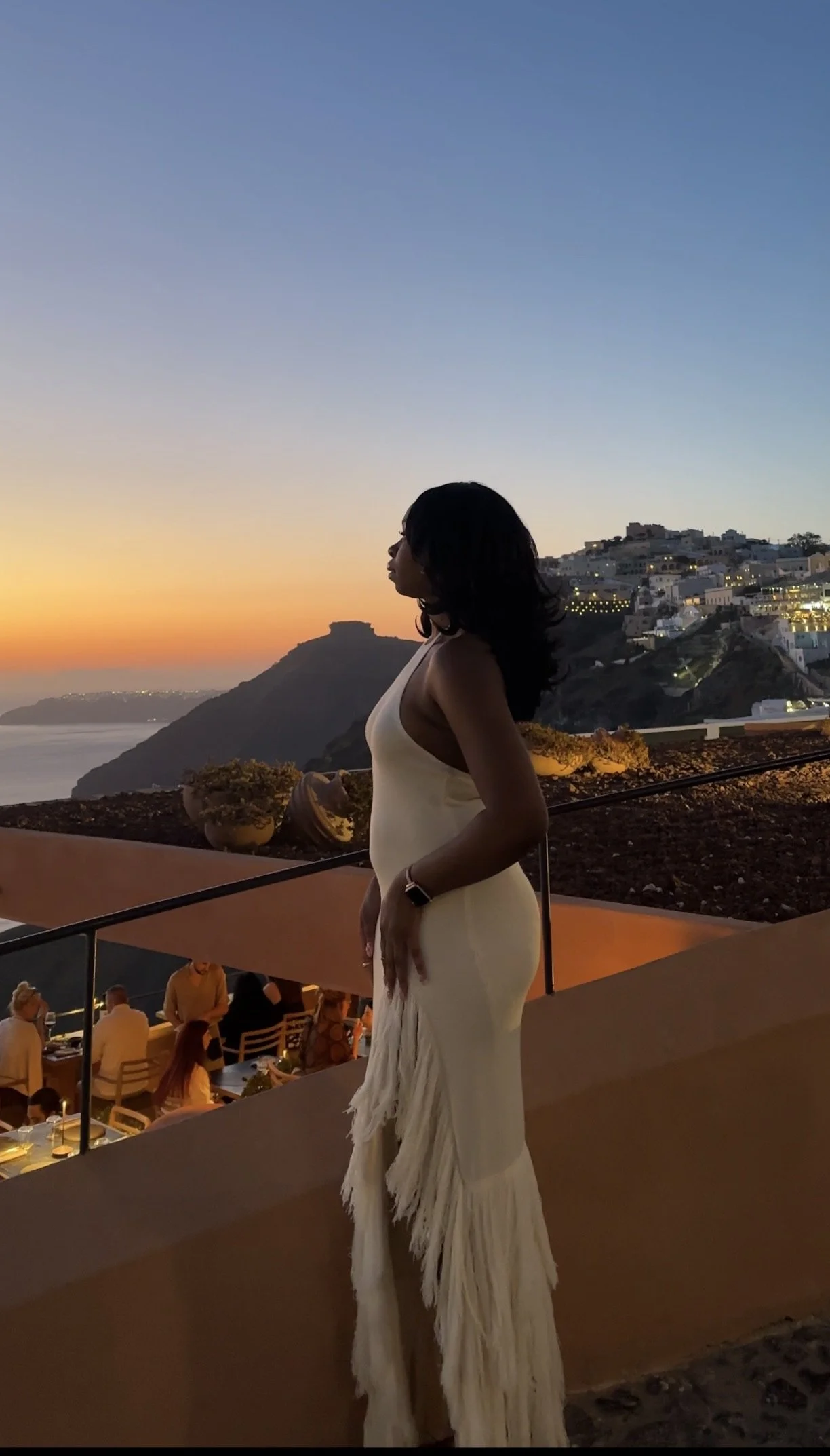 A woman in a white dress standing on an outdoor terrace during sunset with a view of a hillside town in the background.