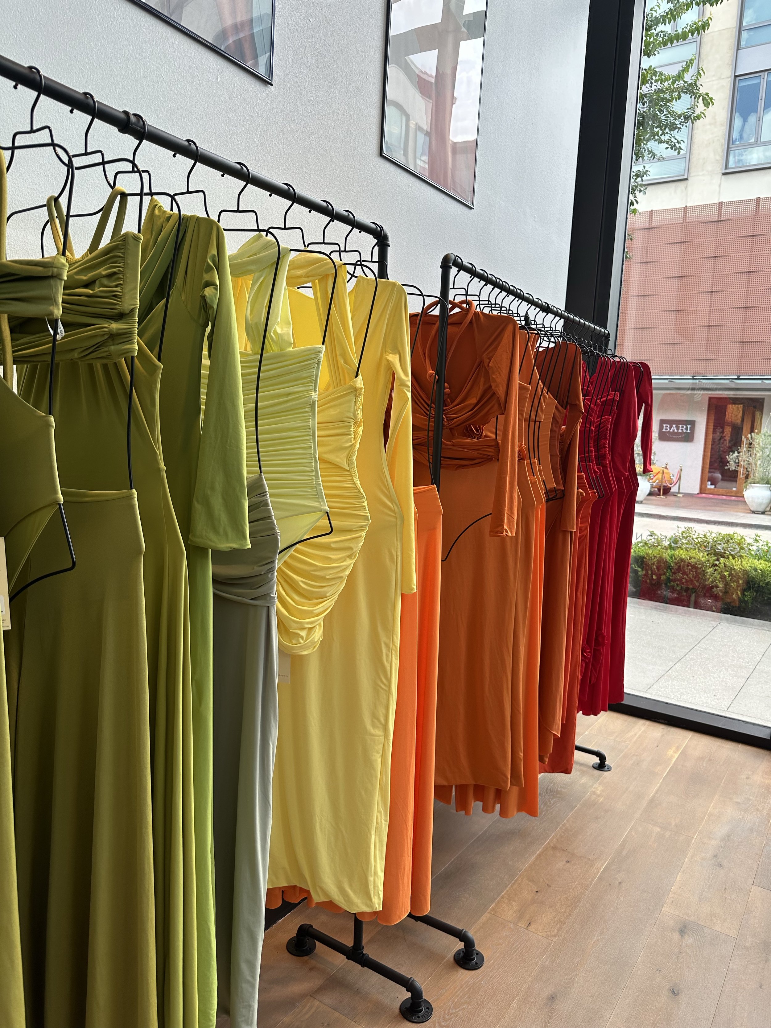 Colorful dresses hanging on a clothing rack near a large window in a store.