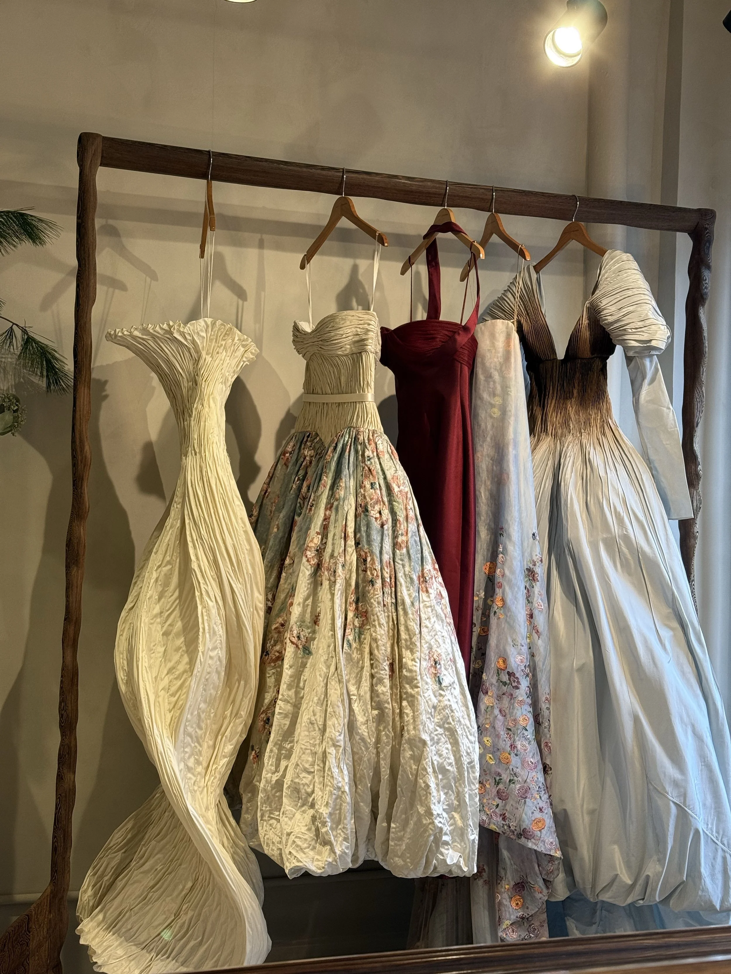Various elegant dresses hanging on wooden hangers on a rustic wooden clothing rack in a softly lit room, mirrored wall reflected behind.