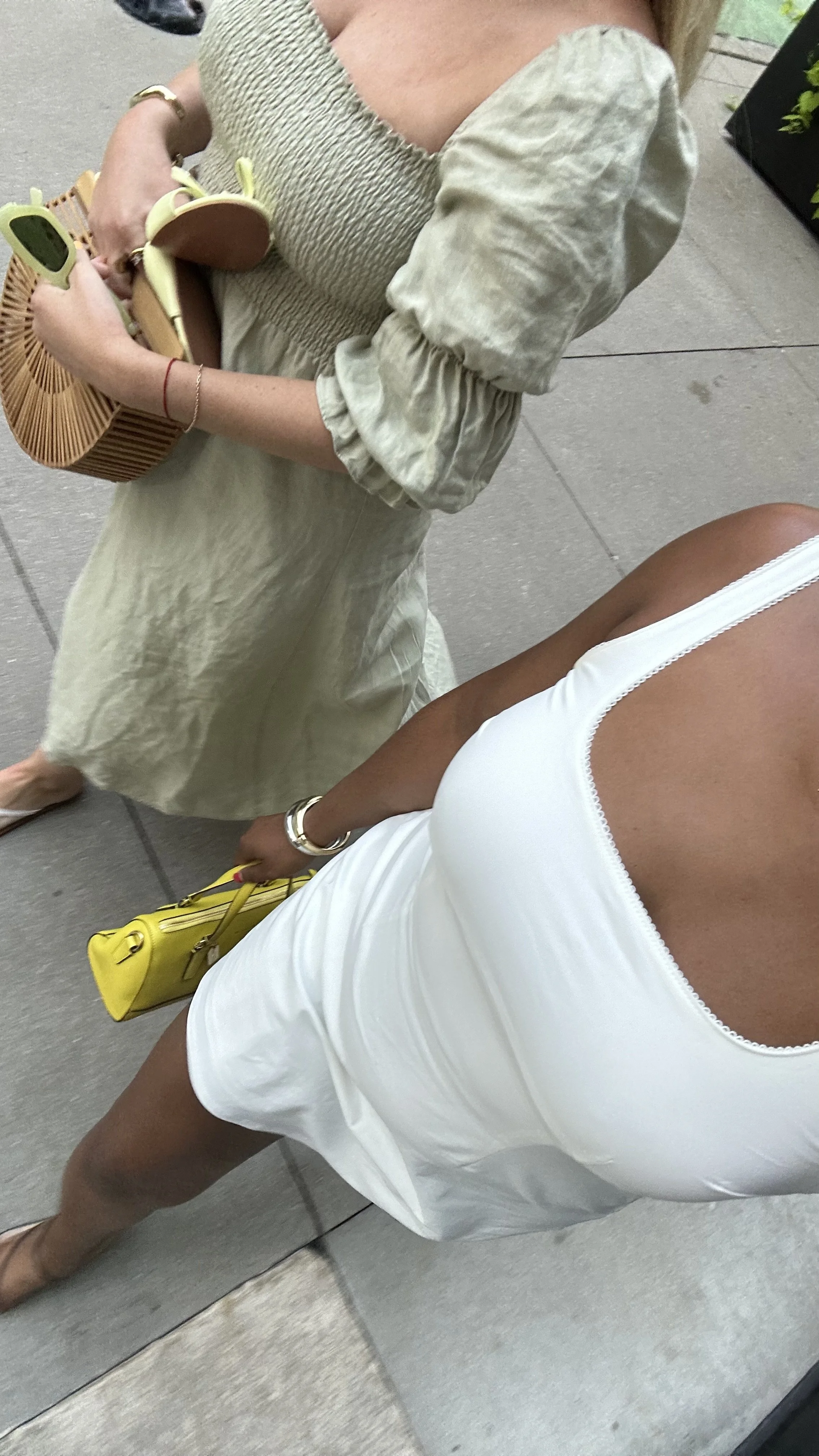A woman wearing a white dress and gold jewelry, holding a yellow handbag, standing in front of a mirror. Another person nearby is holding a tan clutch purse, wearing a beige dress with puffed sleeves, and carrying sunglasses.