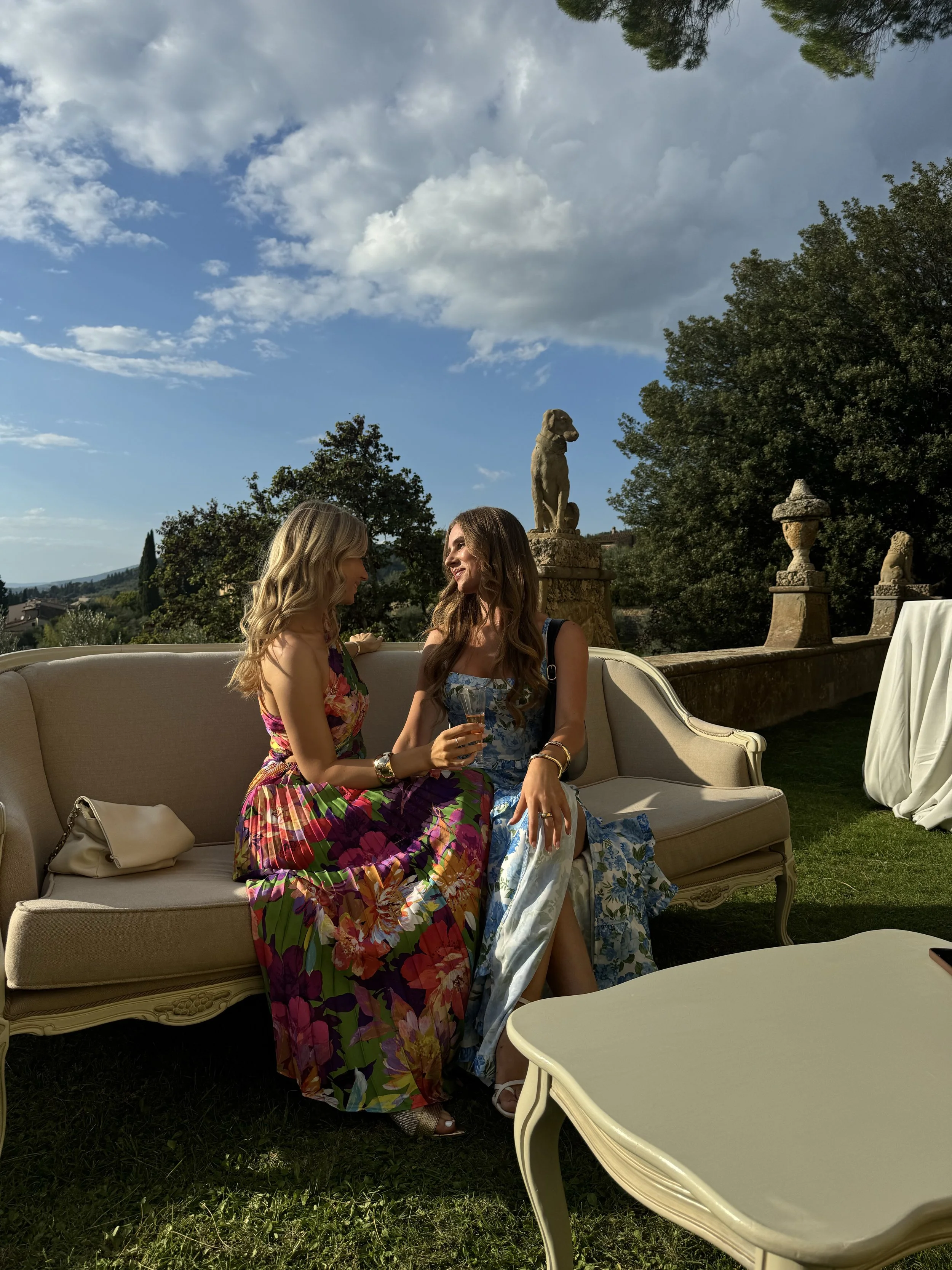Two women sitting on a beige outdoor sofa, smiling and looking at each other, with one holding a glass. They are wearing colorful dresses, and there is a large white table in front of them. Behind them, there are decorative stone sculptures, trees, and a partly cloudy sky.