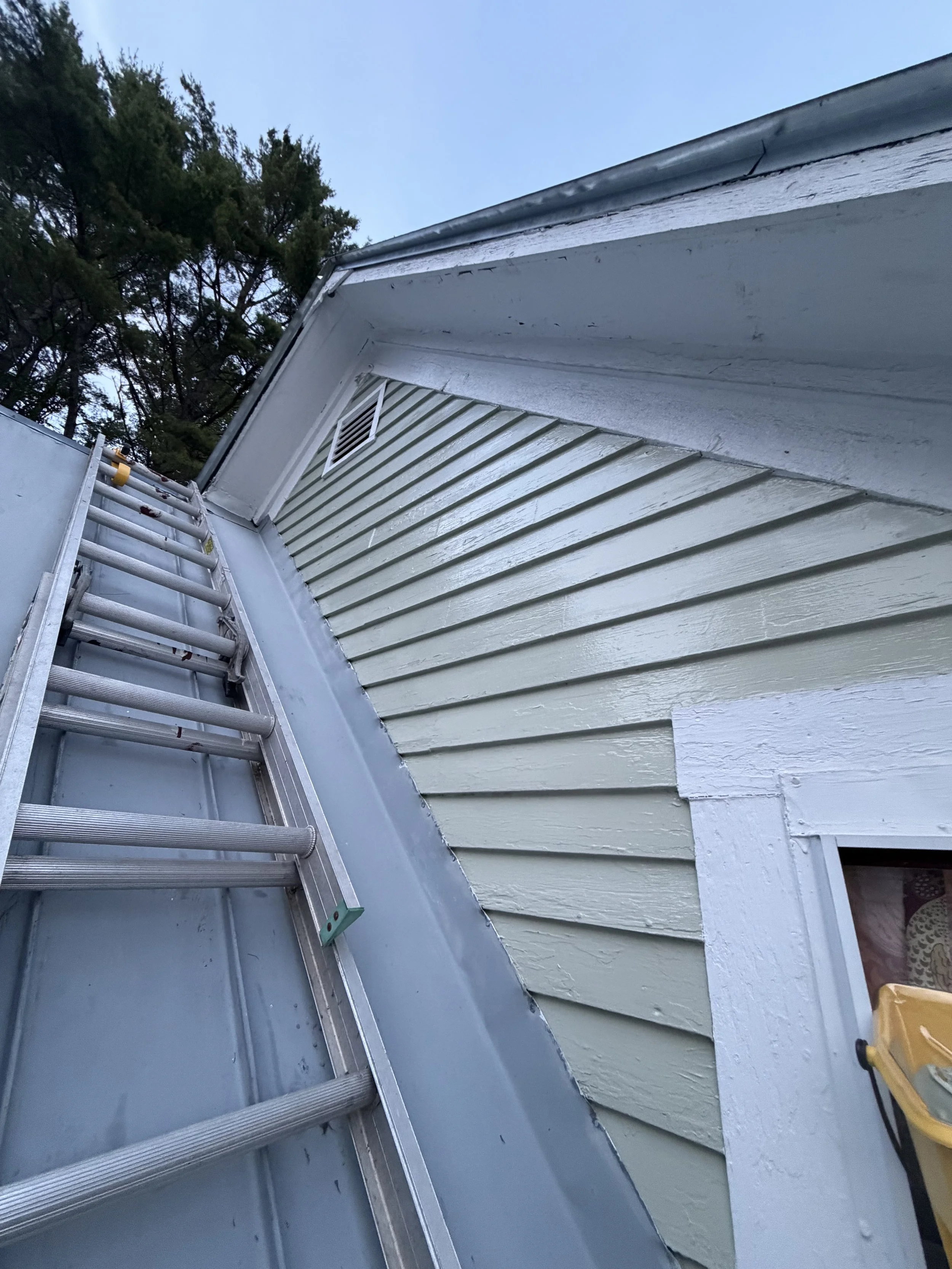An outdoor scene showing the corner of a house with freshly painted wood siding, a metal ladder leaning against the house, a small vent on the siding, and a cloudy sky in the background.