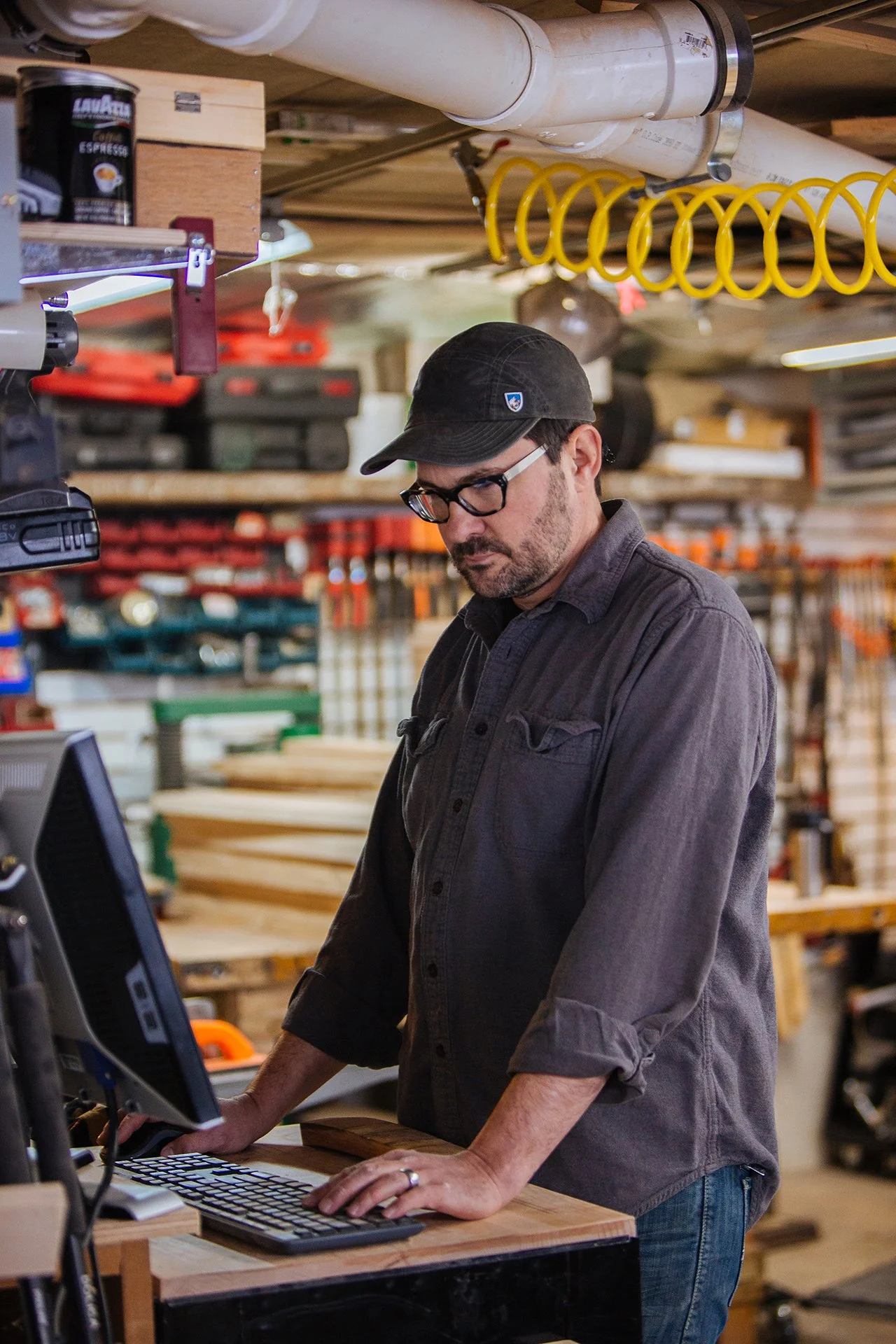A man with glasses wearing a black cap and dark shirt working at a computer in a workshop with woodworking tools and orange extension cords hanging from the ceiling.