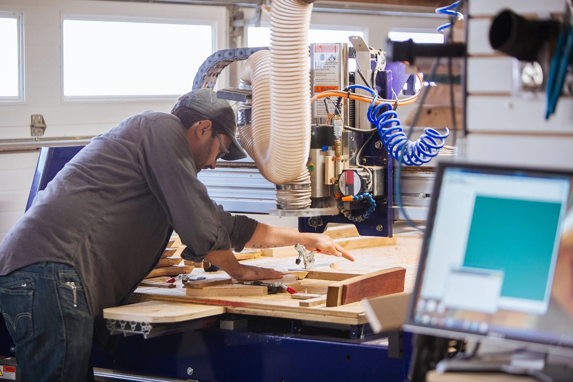 A person working with woodworking equipment at a workbench in a workshop, wearing a gray shirt and baseball cap, with a computer monitor visible in the foreground.