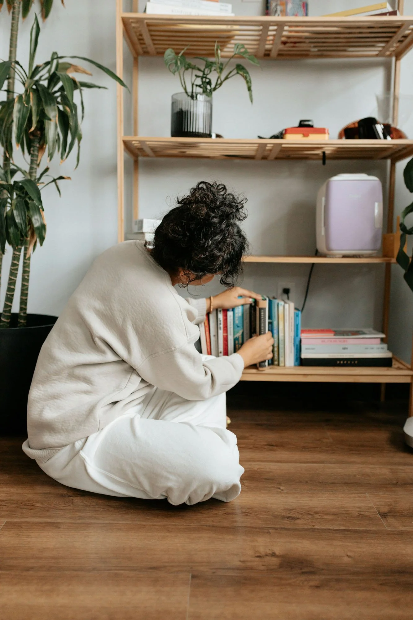 A person with curly hair, dressed in a beige sweatshirt and white pants, is sitting on the wooden floor and looking at a bookshelf.