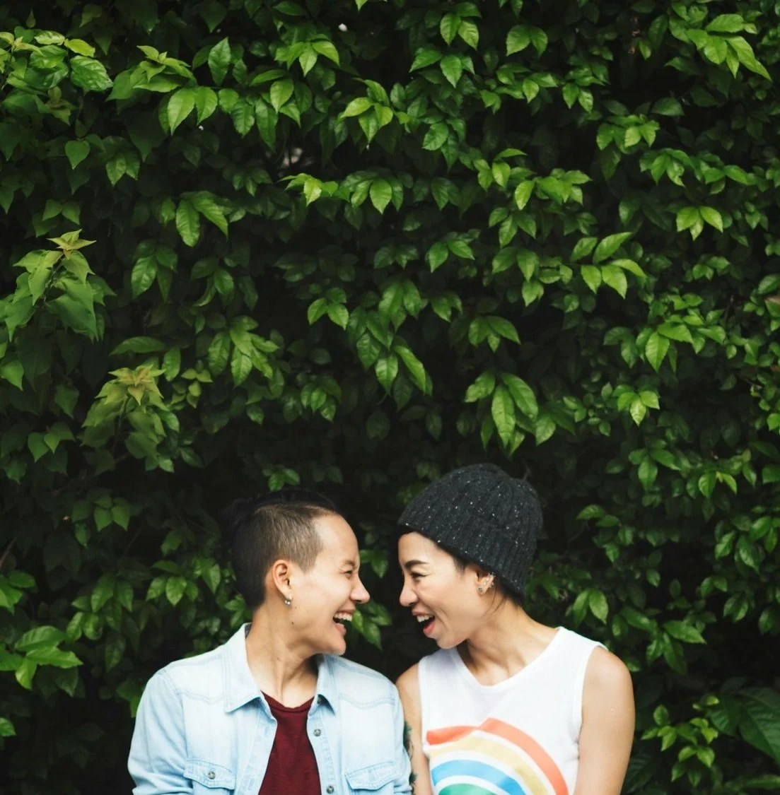 Two women smiling and facing each other in front of lush green foliage.