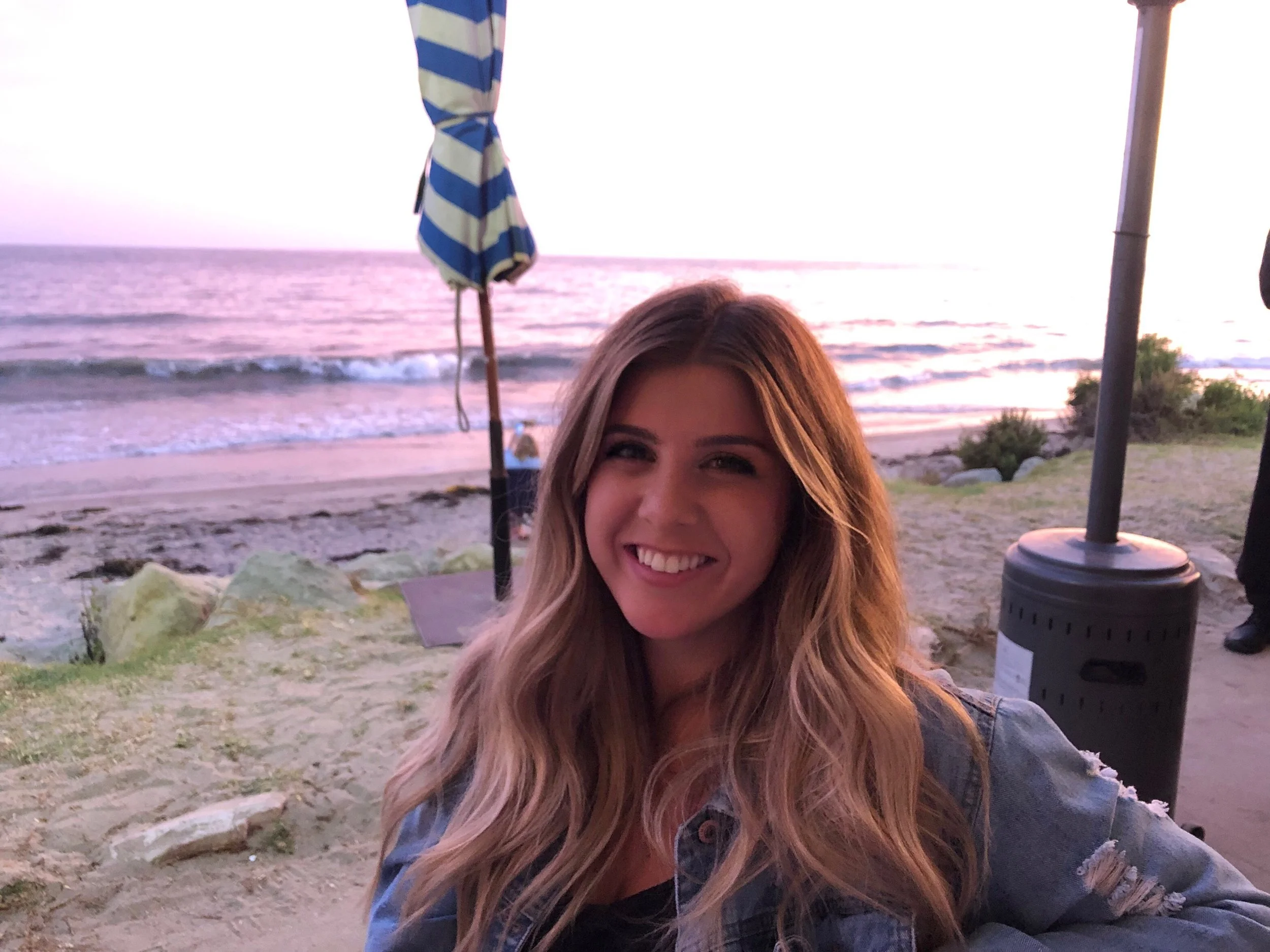 A woman with long blonde hair smiling at the camera, on a beach during sunset. In the background, there is an ocean with small waves, a striped beach umbrella, and some rocks and vegetation near the shoreline.