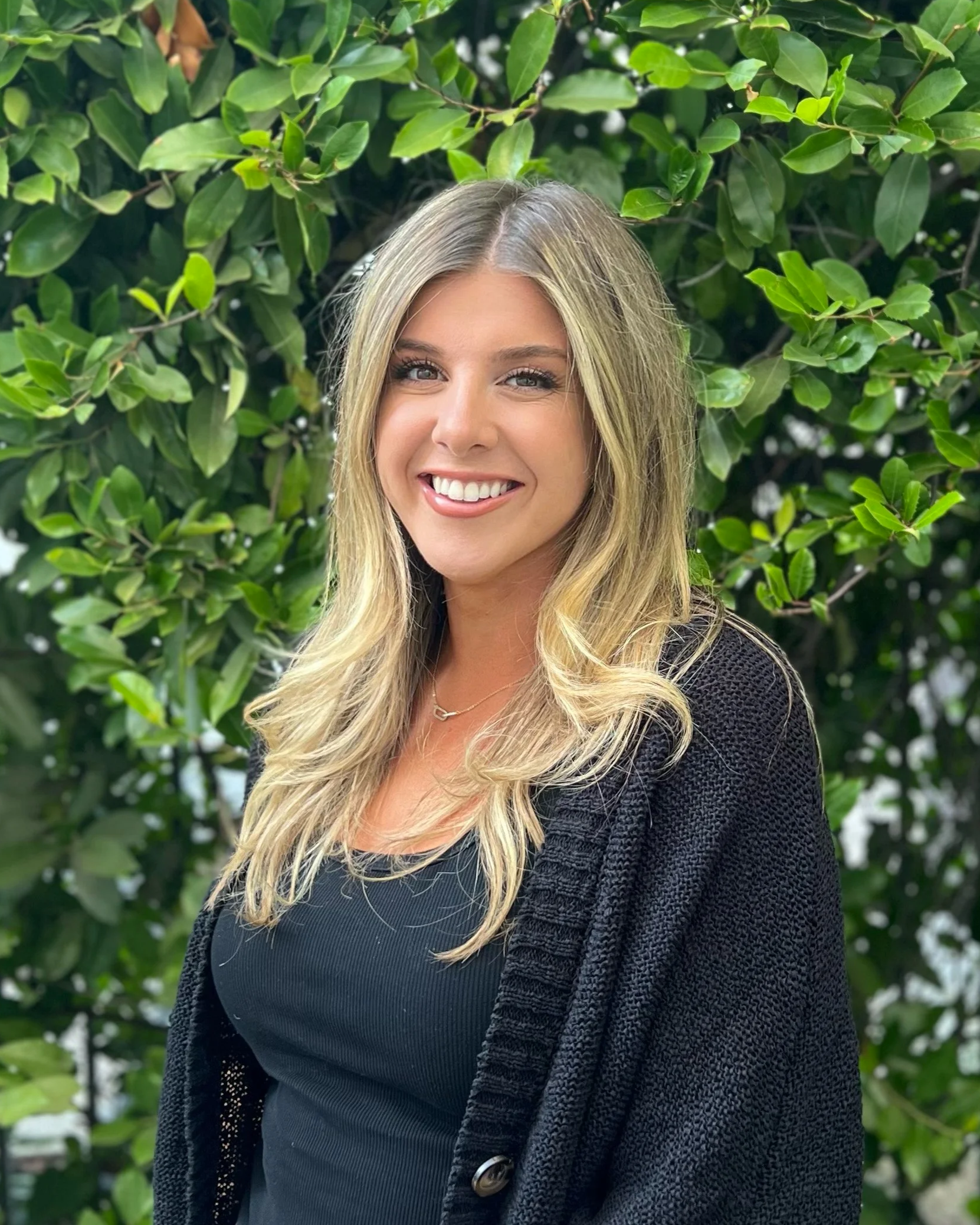 A woman with long blonde hair smiling, wearing a navy top and a black cardigan, standing in front of green leafy bushes.