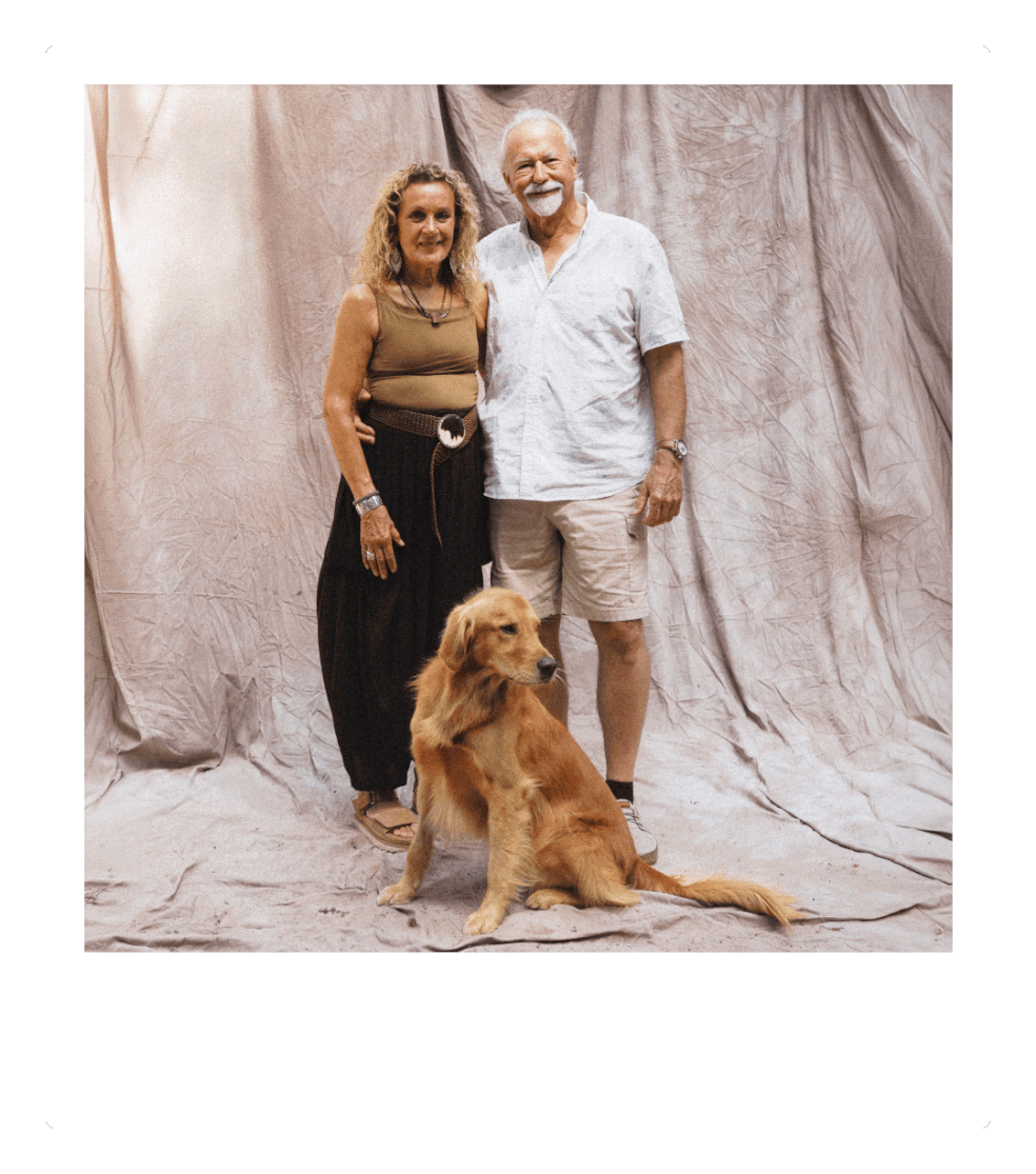 An elderly man and woman standing together indoors against a plain backdrop, with a golden retriever sitting at their feet. The woman has blonde, curly hair and is wearing a sleeveless top and black pants, while the man has white hair and a beard, wearing a white shirt and shorts.