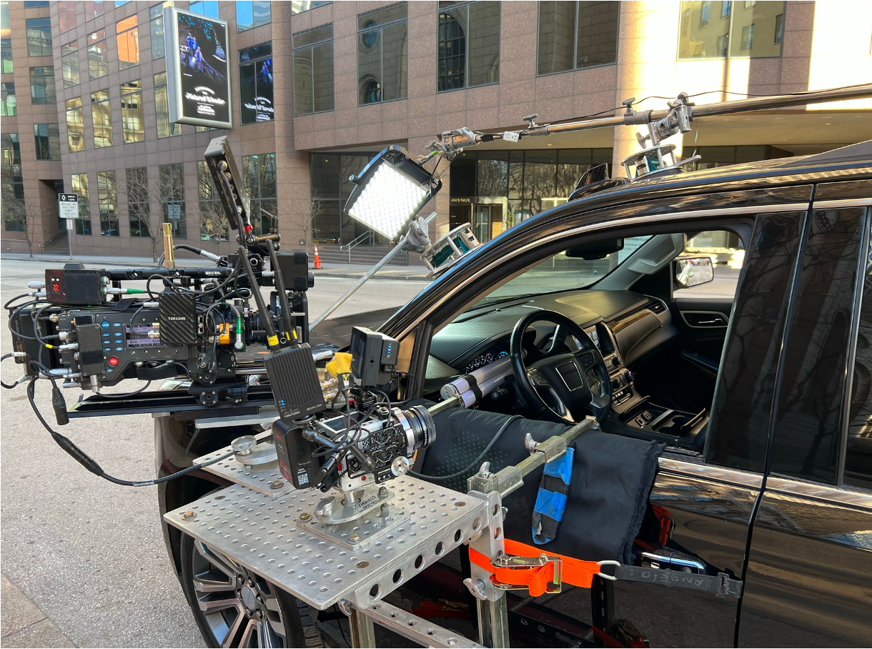 A film or television production camera setup on a sidewalk next to a black SUV, with various equipment and lights, in an urban area with large office buildings.