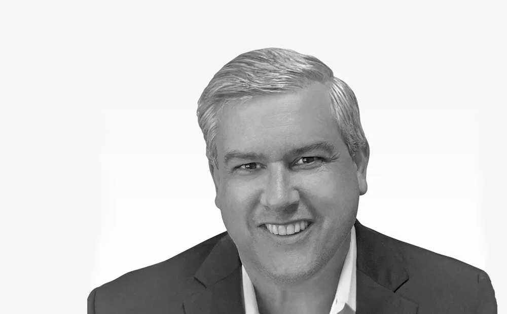 Black and white portrait of J.R. D’Alessandro with blonde hair wearing a suit jacket and white shirt against a plain background.