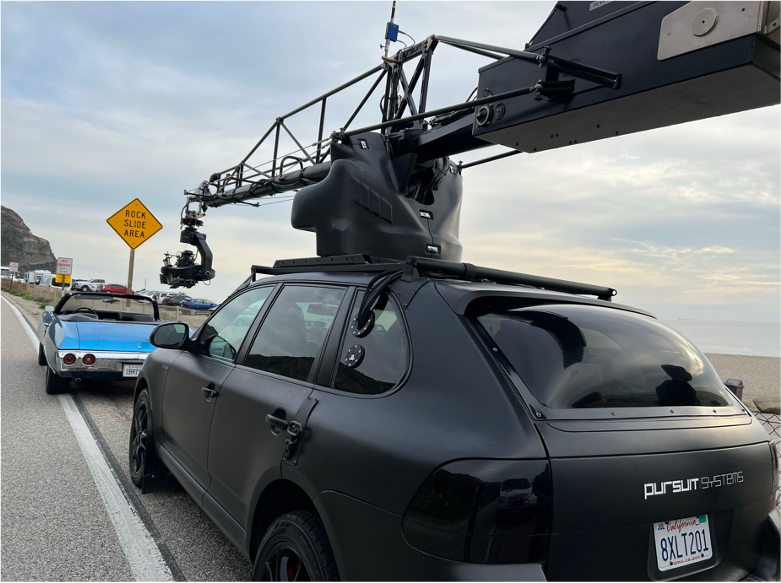 A black Pursuit Systems drone car with a large drone mounted on top, parked on the side of a road near the beach, with a blue classic car behind it and a yellow rock slide warning sign in the background.