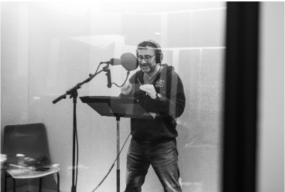 A man wearing glasses, smiling and reading from a paper in a recording studio with a microphone, behind a glass window.