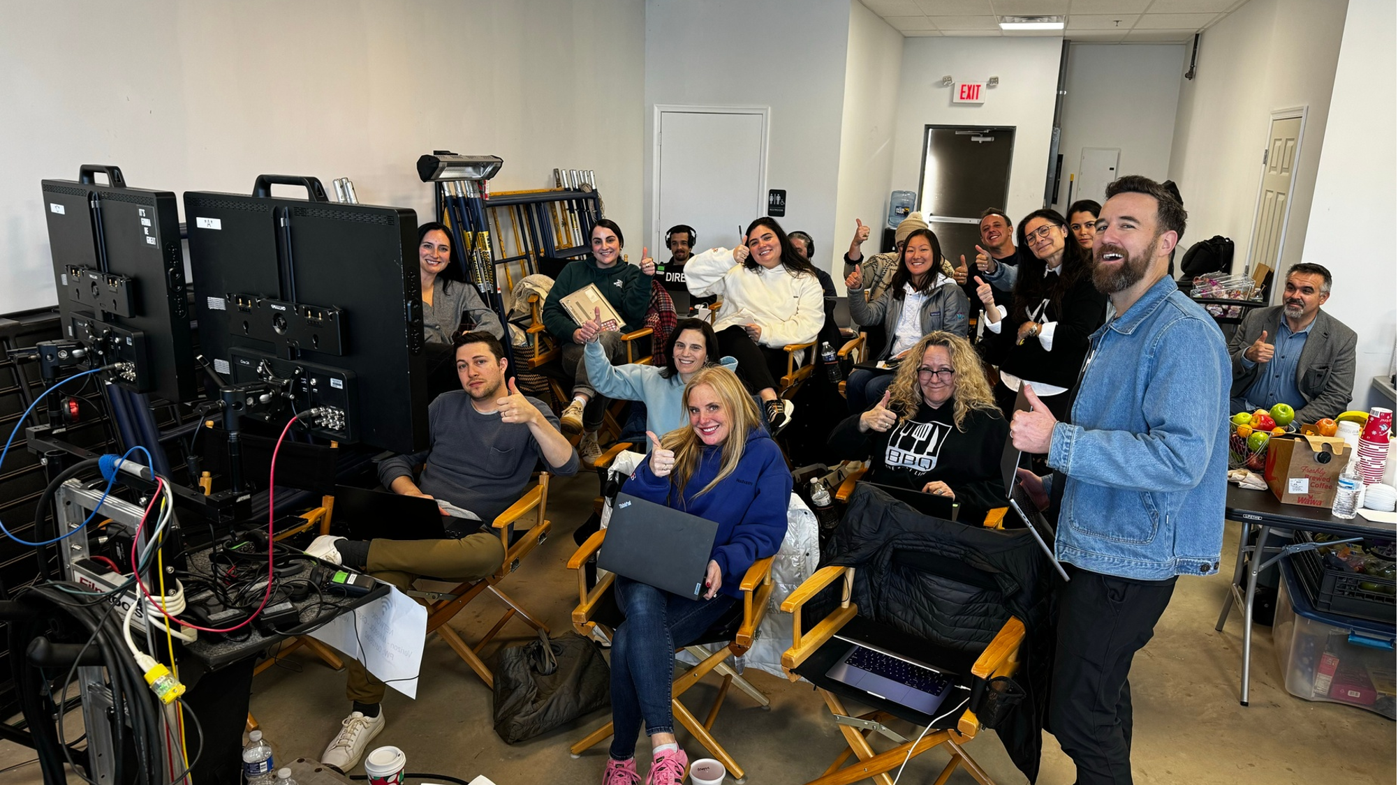 Group of people sitting and standing in a room, smiling and giving thumbs up, with computer monitors and technical equipment in the foreground, and snacks and beverages on a table.