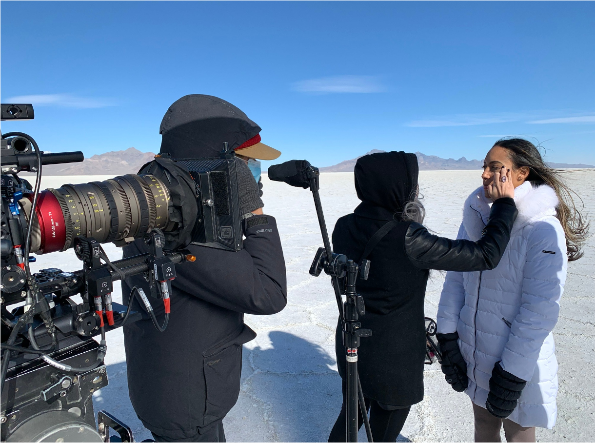 Filming crew members in outdoor desert landscape with salt flats, mountains, and blue sky. One person applies makeup while others operate filming equipment.