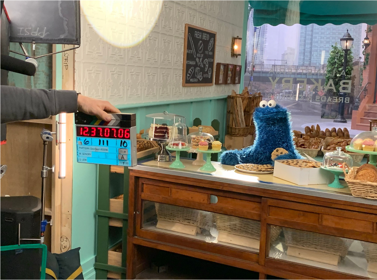 Inside a bakery with a display of baked goods, including cupcakes and cookies. A blue Cookie Monster is on the counter with a cookie in his hand, and a chalkboard sign reads 'Fresh Bread.' Large windows show an urban street scene outside.
