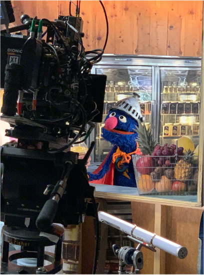 Behind-the-scenes of a TV set with a puppet character wearing a white helmet, blue fur, orange scarf, and large pink nose, standing in front of a display case of fresh fruits and vegetables.