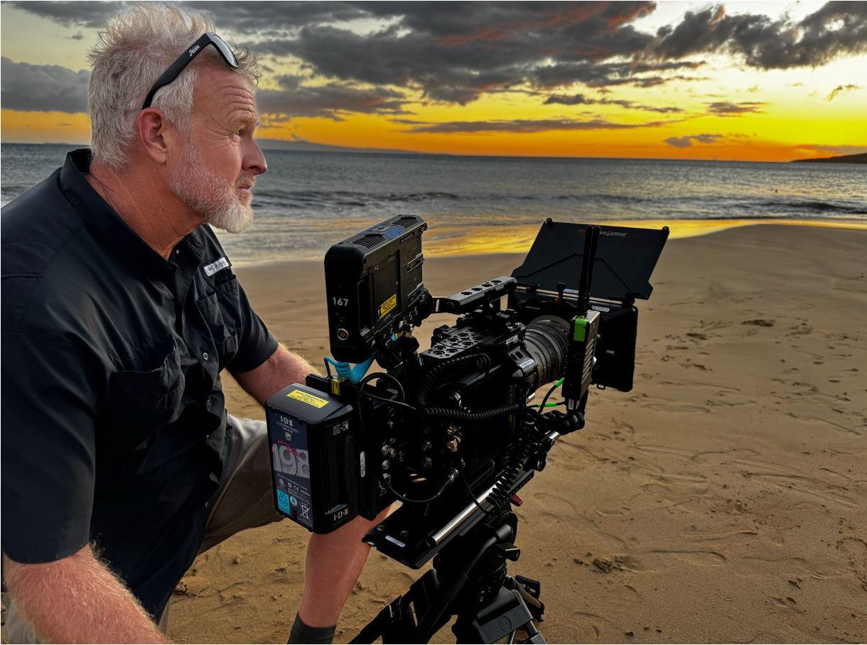 An older man with gray hair and beard operating a professional camera on a sandy beach at sunset, with the ocean and a sky filled with clouds in the background.