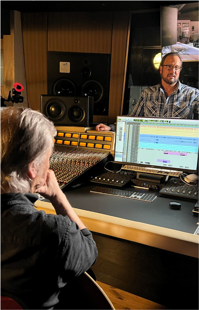 In a recording studio, a man with gray hair and a black shirt sits in front of a mixing console, looking at a computer screen displaying audio editing software. Another man with glasses and a plaid shirt stands behind him, observing the work. Large studio monitors and wooden paneling are visible in the background.