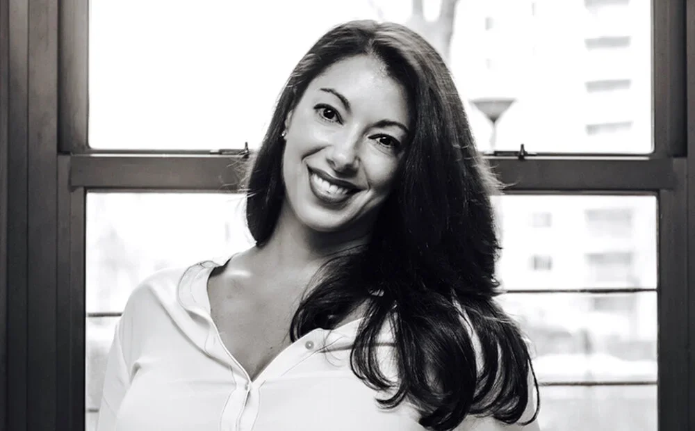 Black and white photo of Coco Seijas with long dark hair, wearing a white shirt, standing in front of a window.