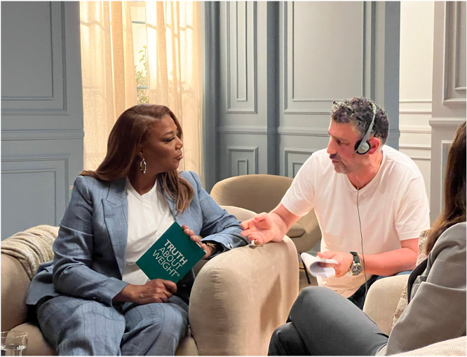 A woman in a blue plaid suit is sitting on a beige sofa, holding a book titled 'Truth About Weight', while Rob Farber in a white t-shirt with interview headphones talks to her and gestures with his hand.