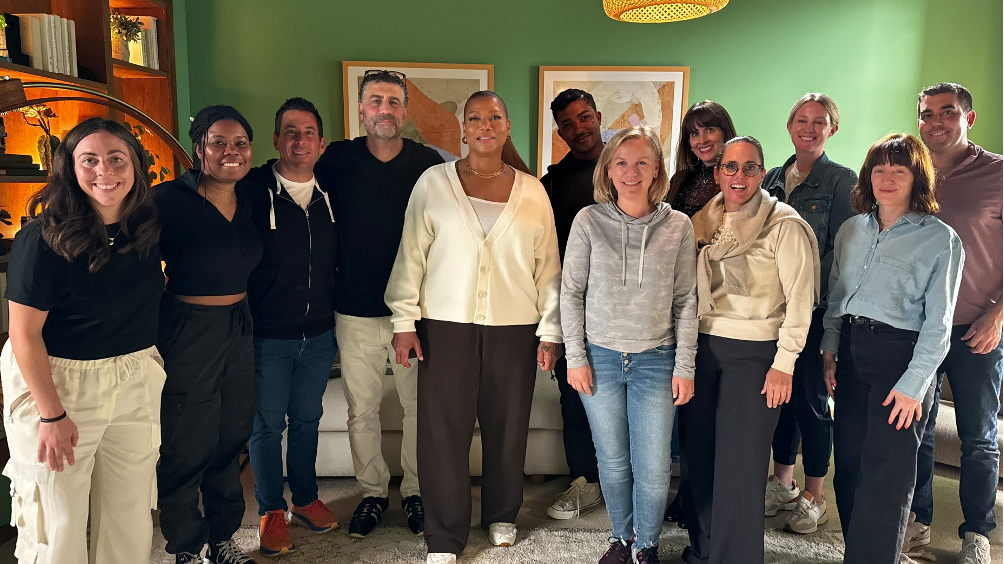 A group of fifteen diverse people standing together indoors, smiling, with a green wall, artwork, and bookshelves in the background.