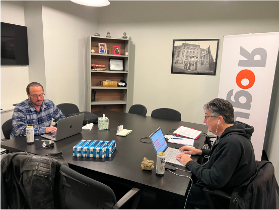 Two men sitting at a conference table working on laptops in a meeting room, with a bookshelf, framed picture, and banner in the background.