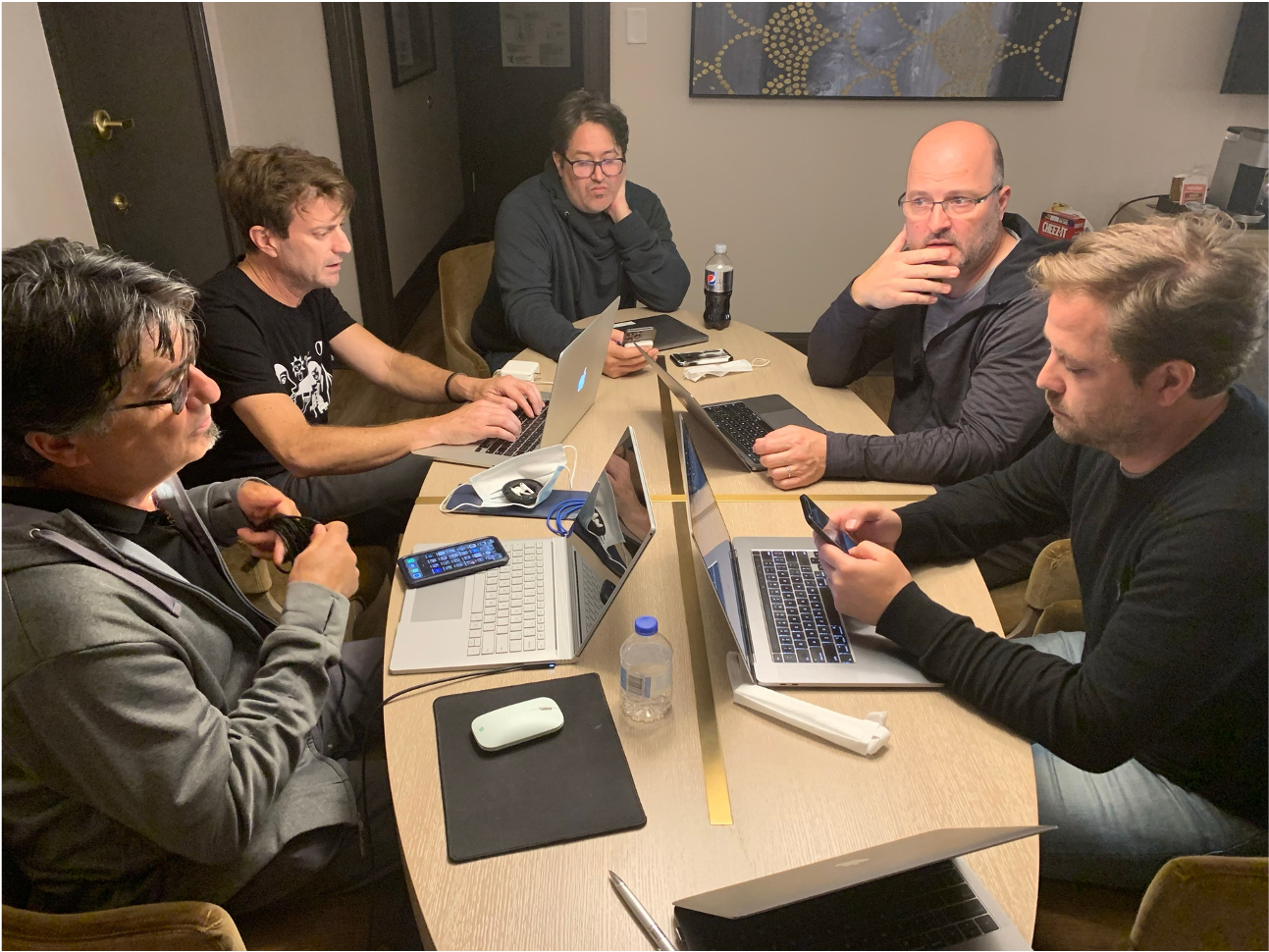 Six men are sitting around a rectangular table with laptops, smartphones, and water bottles, engaged in a discussion or meeting in a room with framed artwork on the wall.