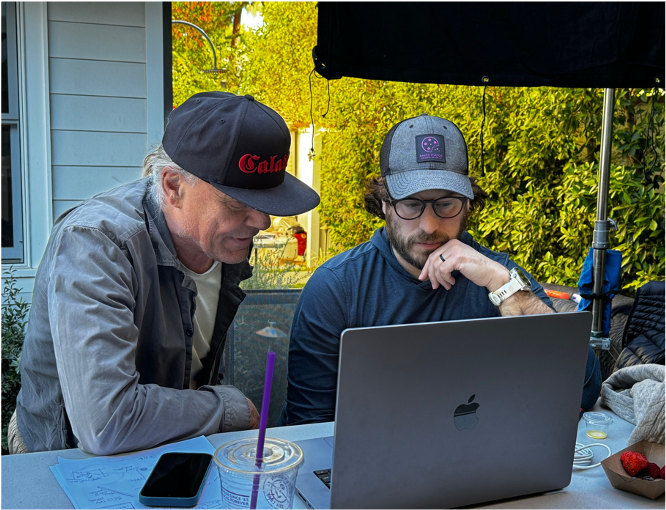 Two men sitting at a table outdoors, looking at a laptop, with one wearing a black cap with red writing and the other wearing a gray cap and glasses.
