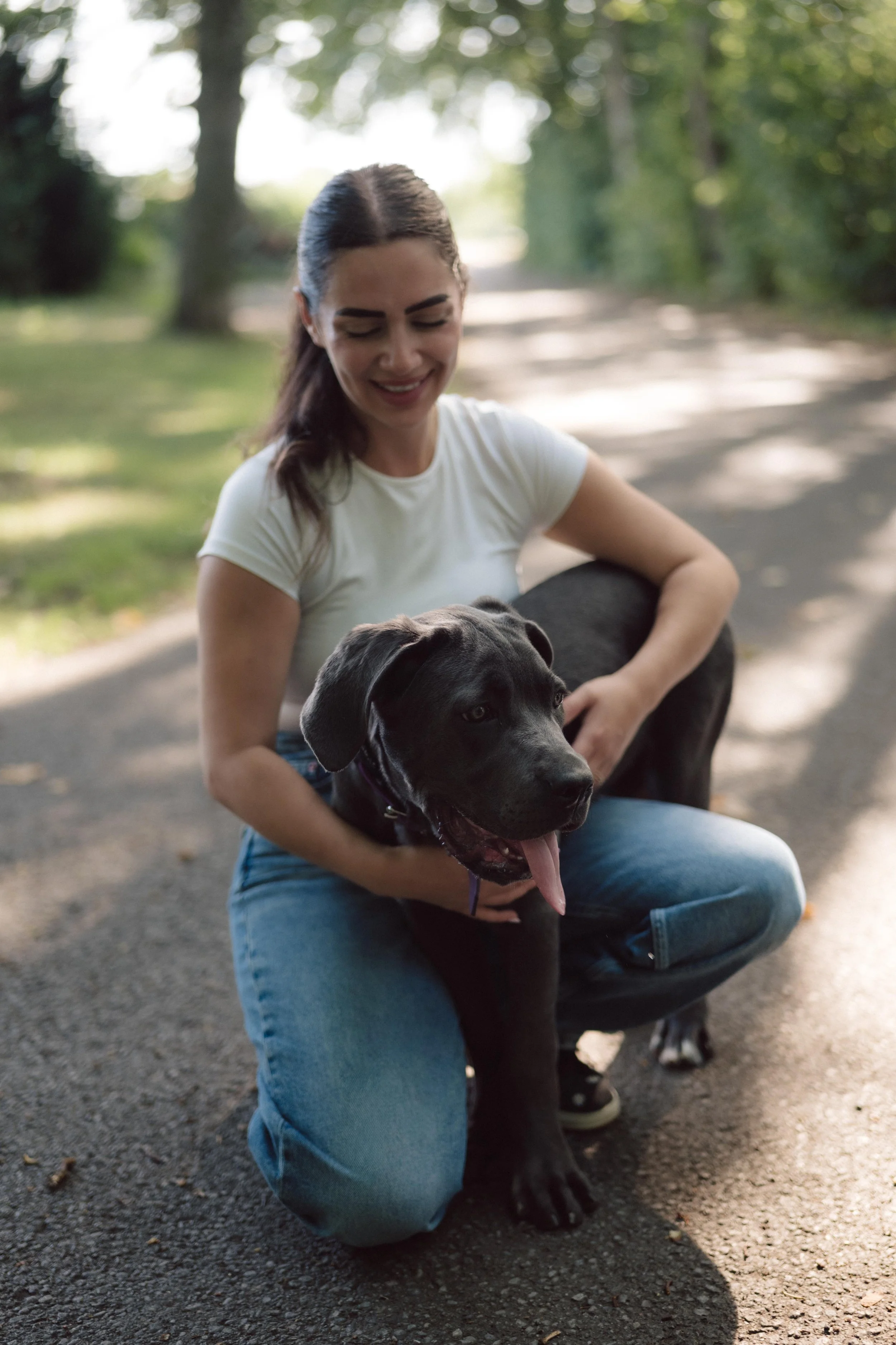 Hundetrainerin Natascha von Hamrah Hundetraining kniet lächelnd neben einem schwarzen Hund auf einem Waldweg