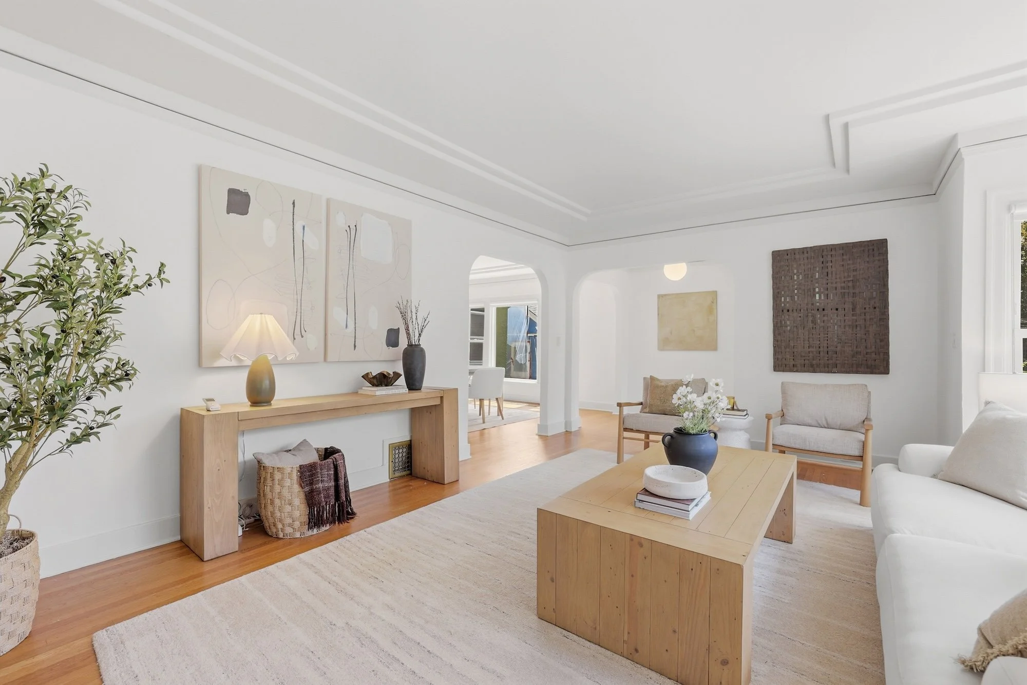 A living room with white walls and wood flooring, decorated with abstract artwork, plants, and minimalist furniture including a wooden bench, coffee table, and sofas.