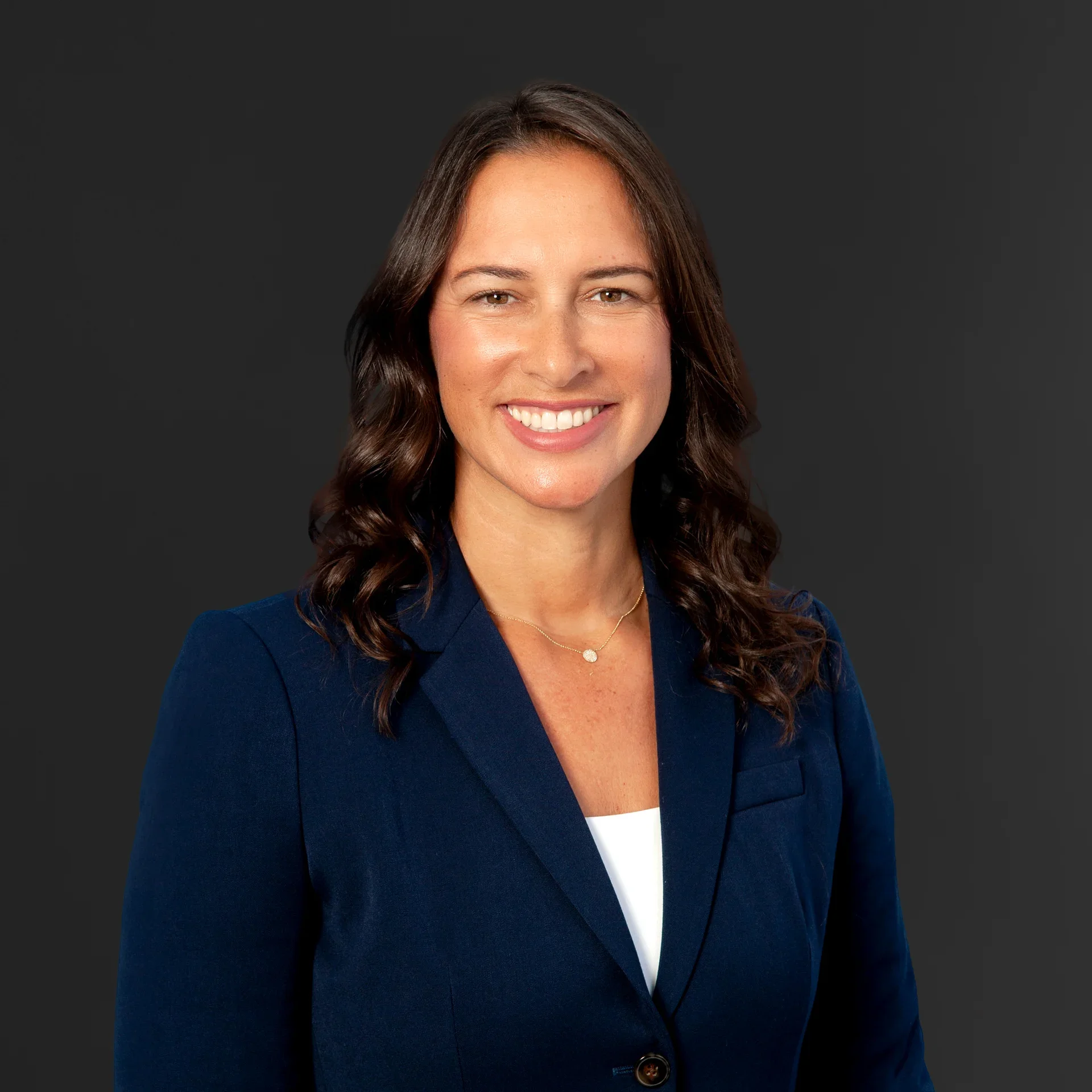 Close-up of a woman with brown hair, smiling, wearing a navy blazer and a white shirt, against a dark gray background.