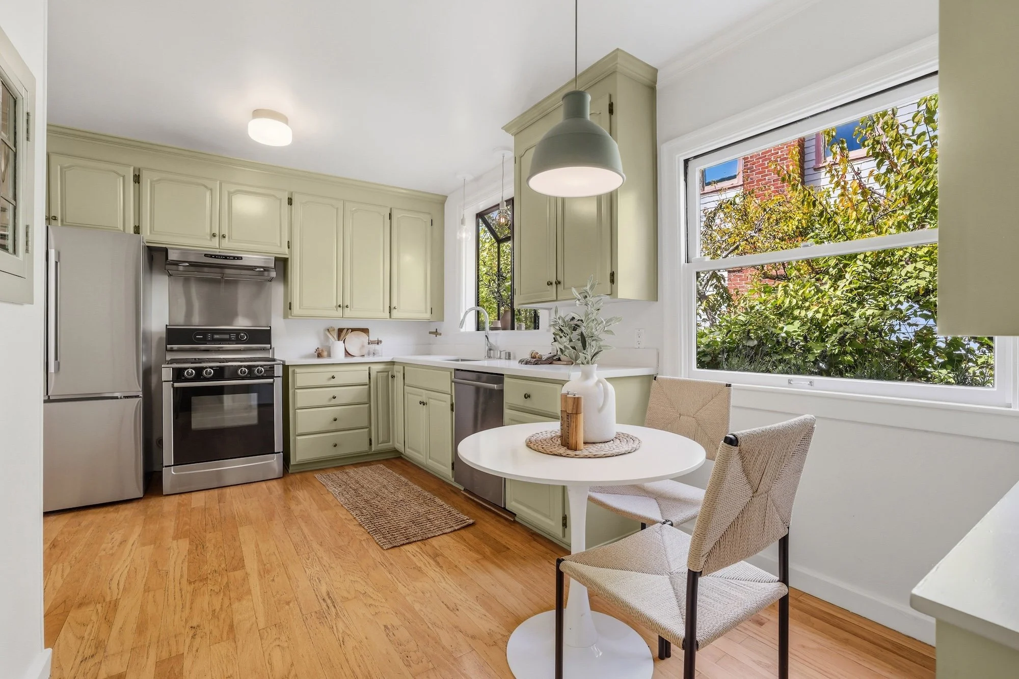 Kitchen with cream-colored cabinets, stainless steel appliances, a round white table with chairs, large window showing greenery outside, and pendant lighting.