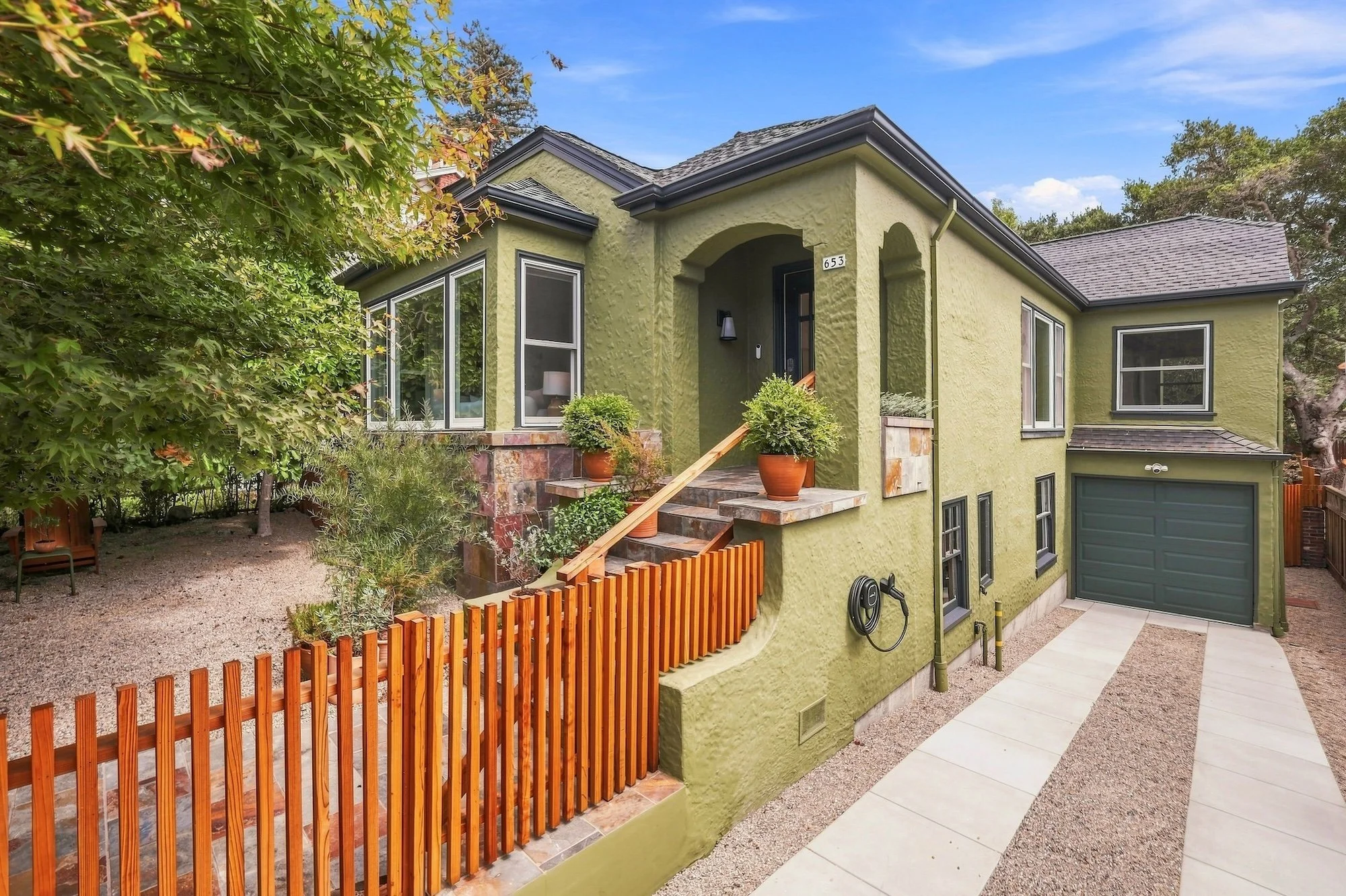 A modern green multi-story house with a garage, surrounded by a wooden fence, plants, and trees, under a blue sky.