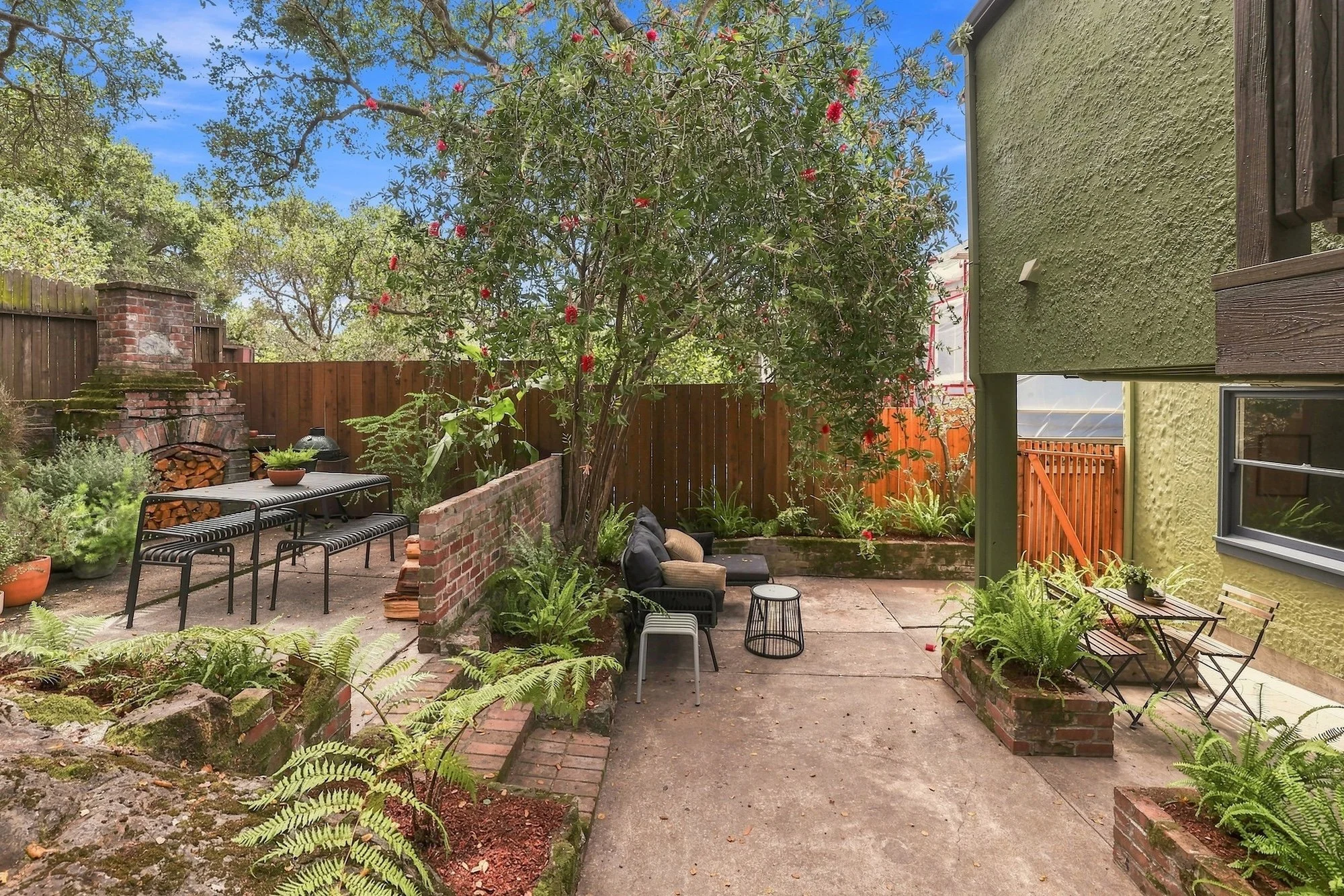 A backyard patio with outdoor furniture, lush greenery, potted plants, a wood fire grill, and a wooden fence under a clear blue sky.
