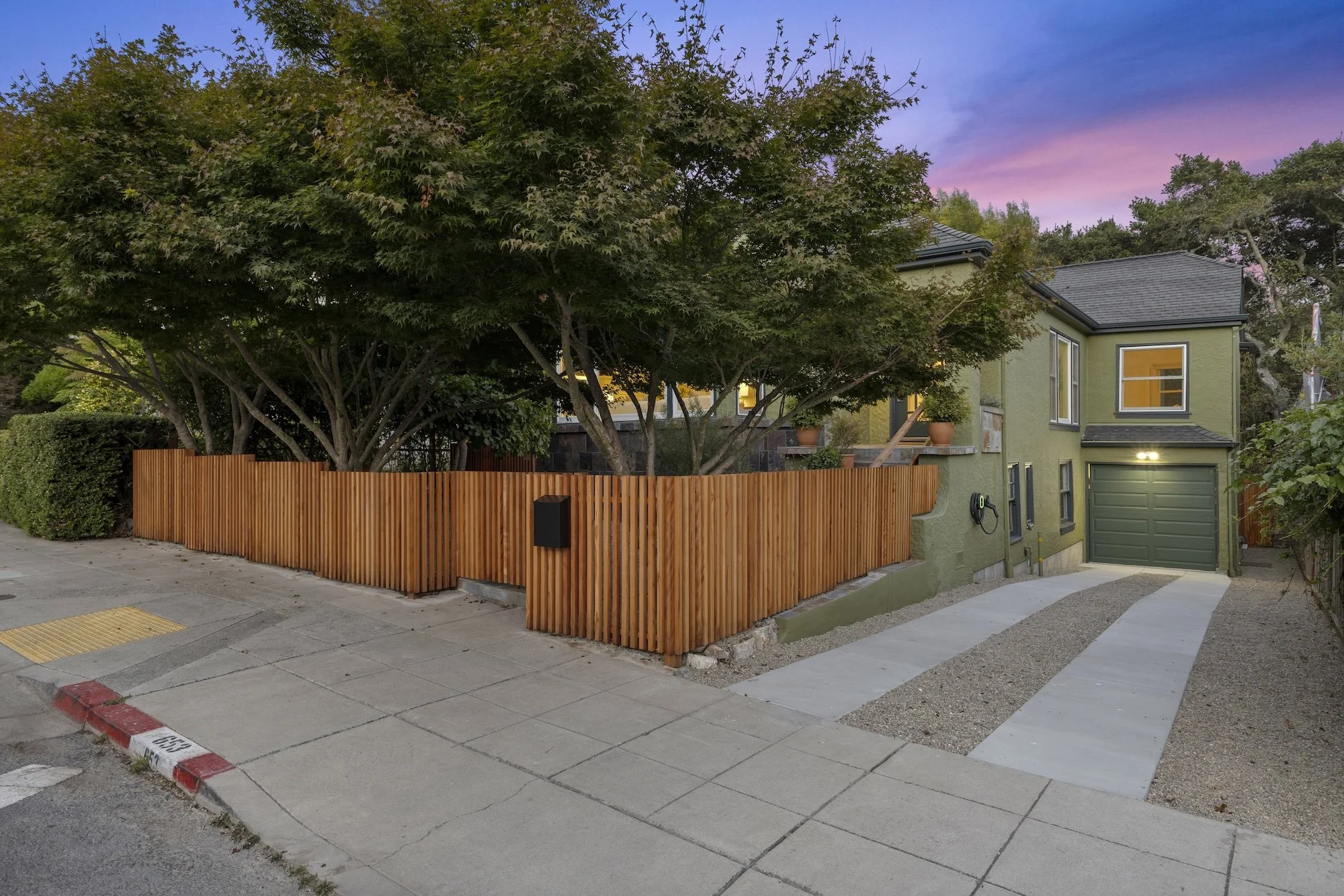 A residential house with a yellowish-green exterior and a dark gray roof, partially obscured by a large tree with dense green foliage. The house has potted plants on the balcony and a garage door at the front. The scene is during dusk with a purple a
