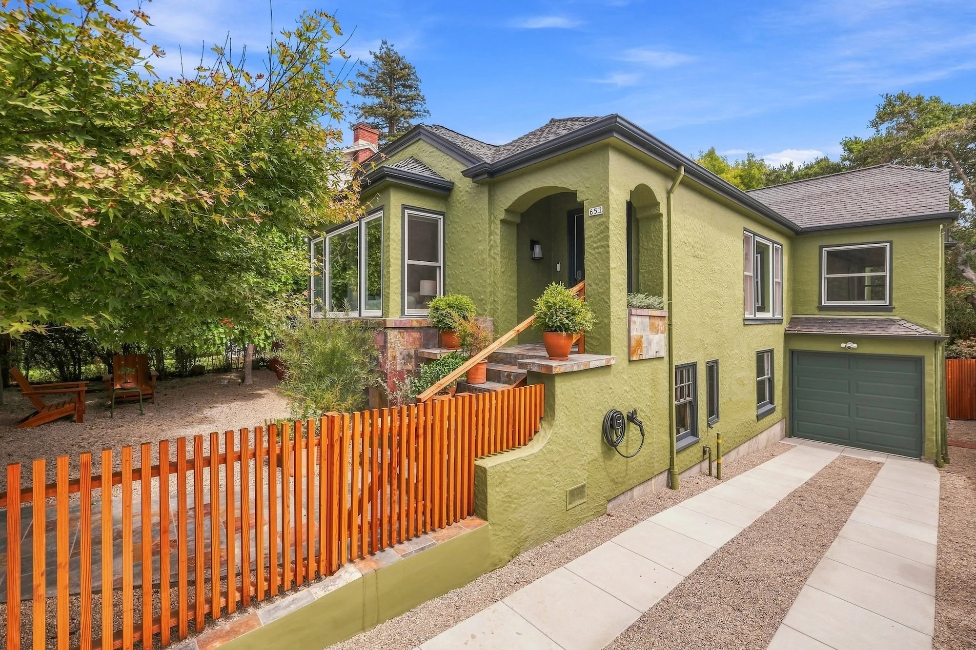 A green two-story house with large windows, a sloped roof, and a small front porch with potted plants. There is a wooden fence and trees surrounding the house, with a gravel area, a paved pathway, and a blue sky with few clouds.