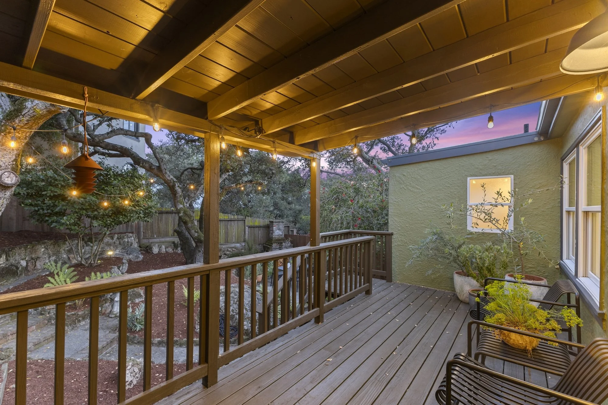 Wood deck with railing under a ceiling, string lights hanging, potted plants, trees, and a sunset sky in the background.