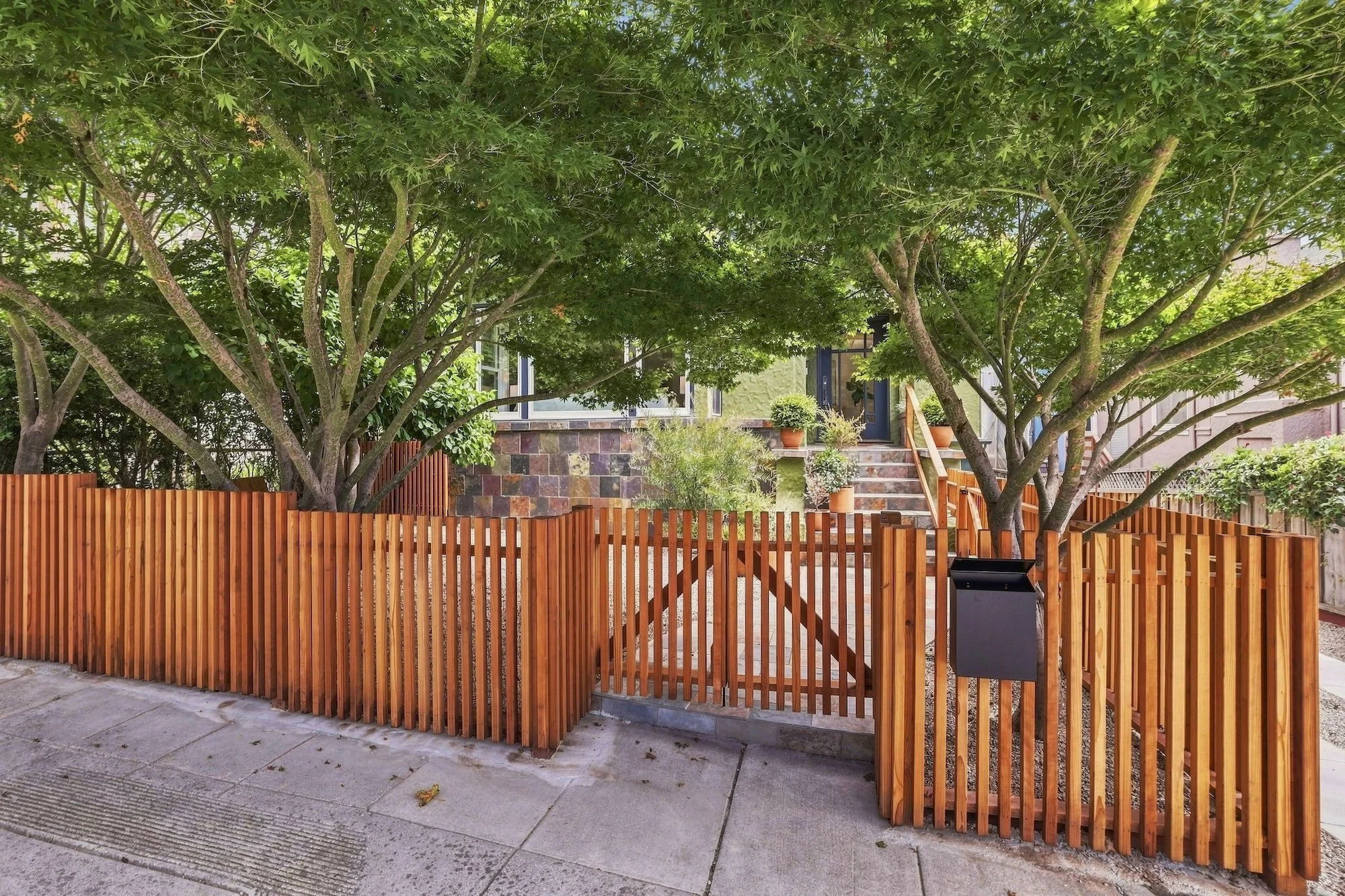 Front yard of a house with a wooden fence, trees with green leaves, potted plants on stairs, and a house with a porch and steps.