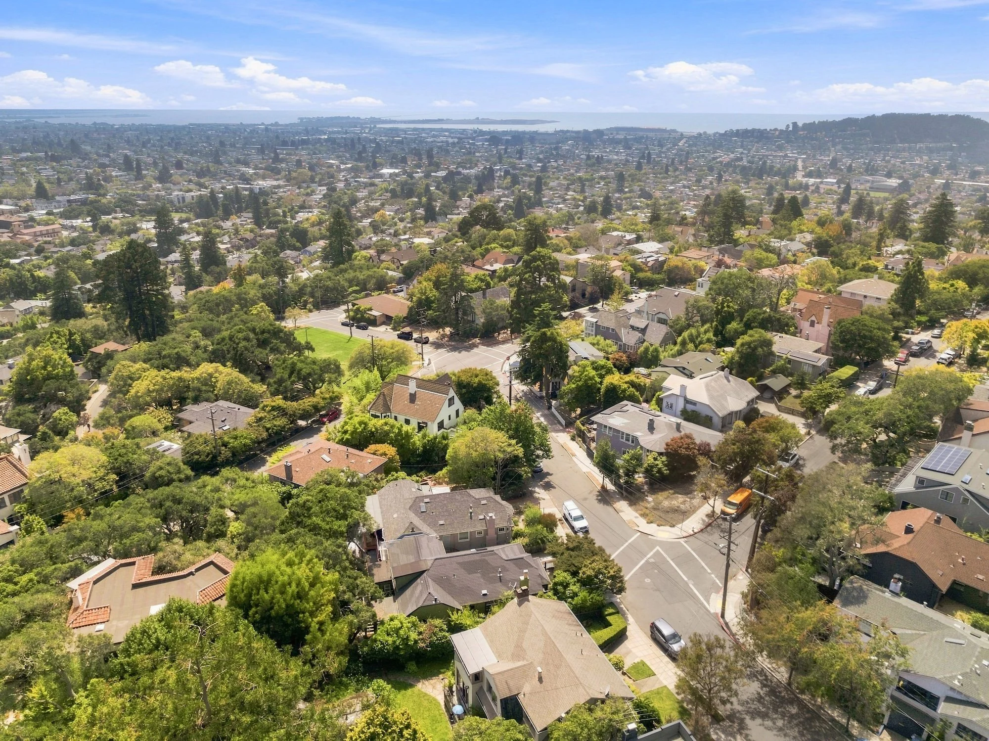 Aerial view of a suburban neighborhood with houses, trees, and streets, with the ocean in the background under a blue sky with some clouds.