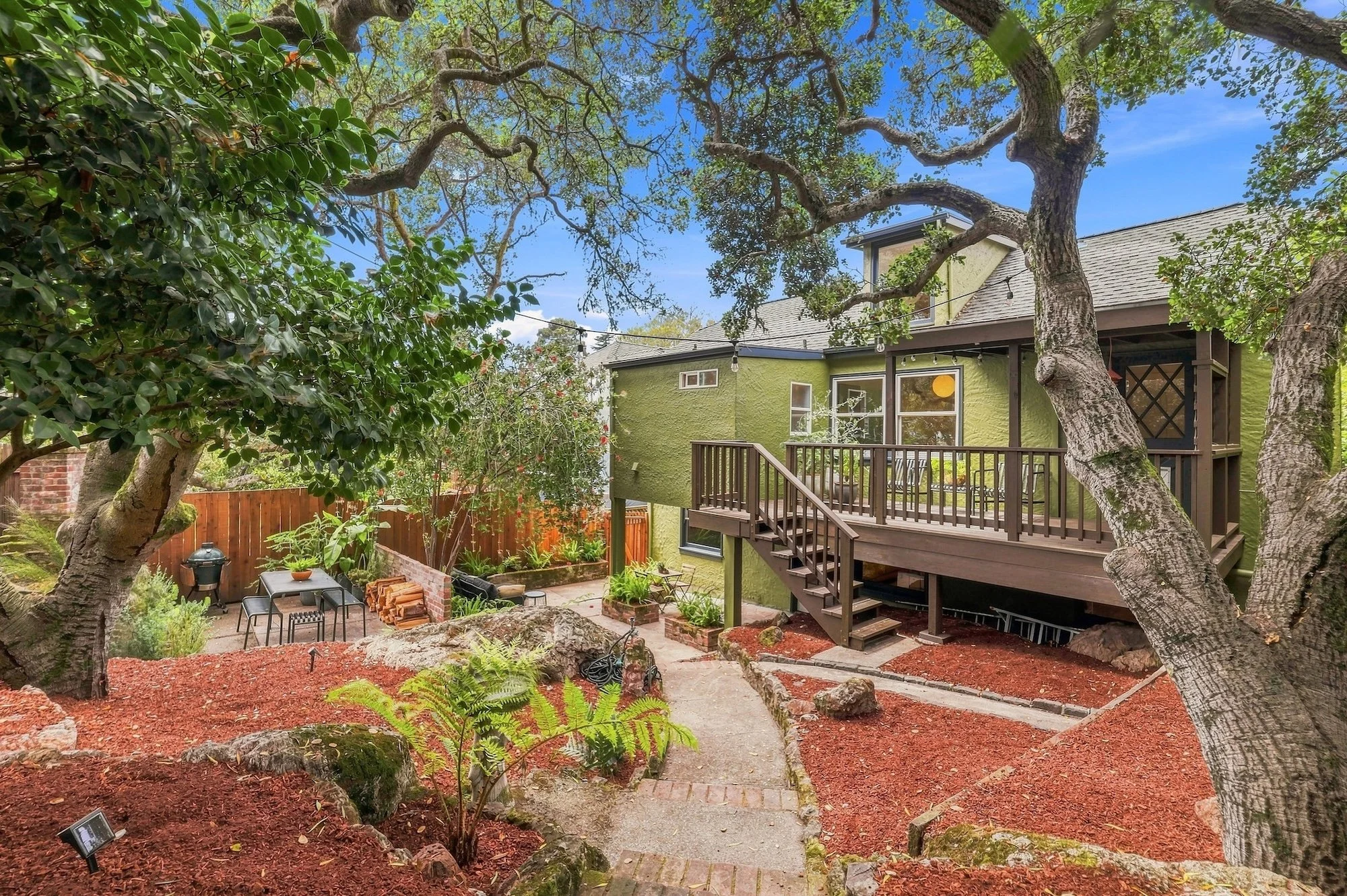 A backyard garden with large trees, a raised deck attached to a green house, patio furniture, and a brick-lined garden area with mulch, plants, and a small fire pit.