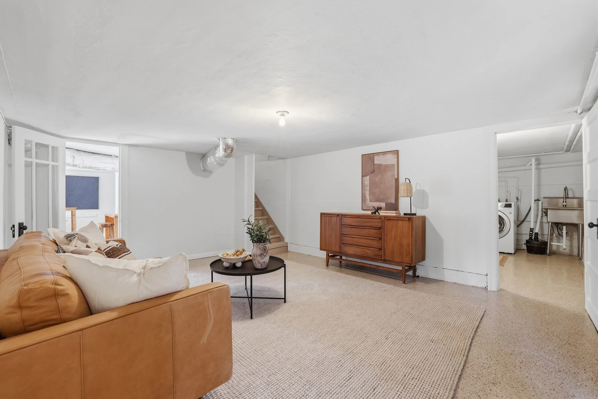 Living room with beige sofa, wooden dresser, artwork, and a round table with a vase of greenery, carpeted floor, and visible laundry area.