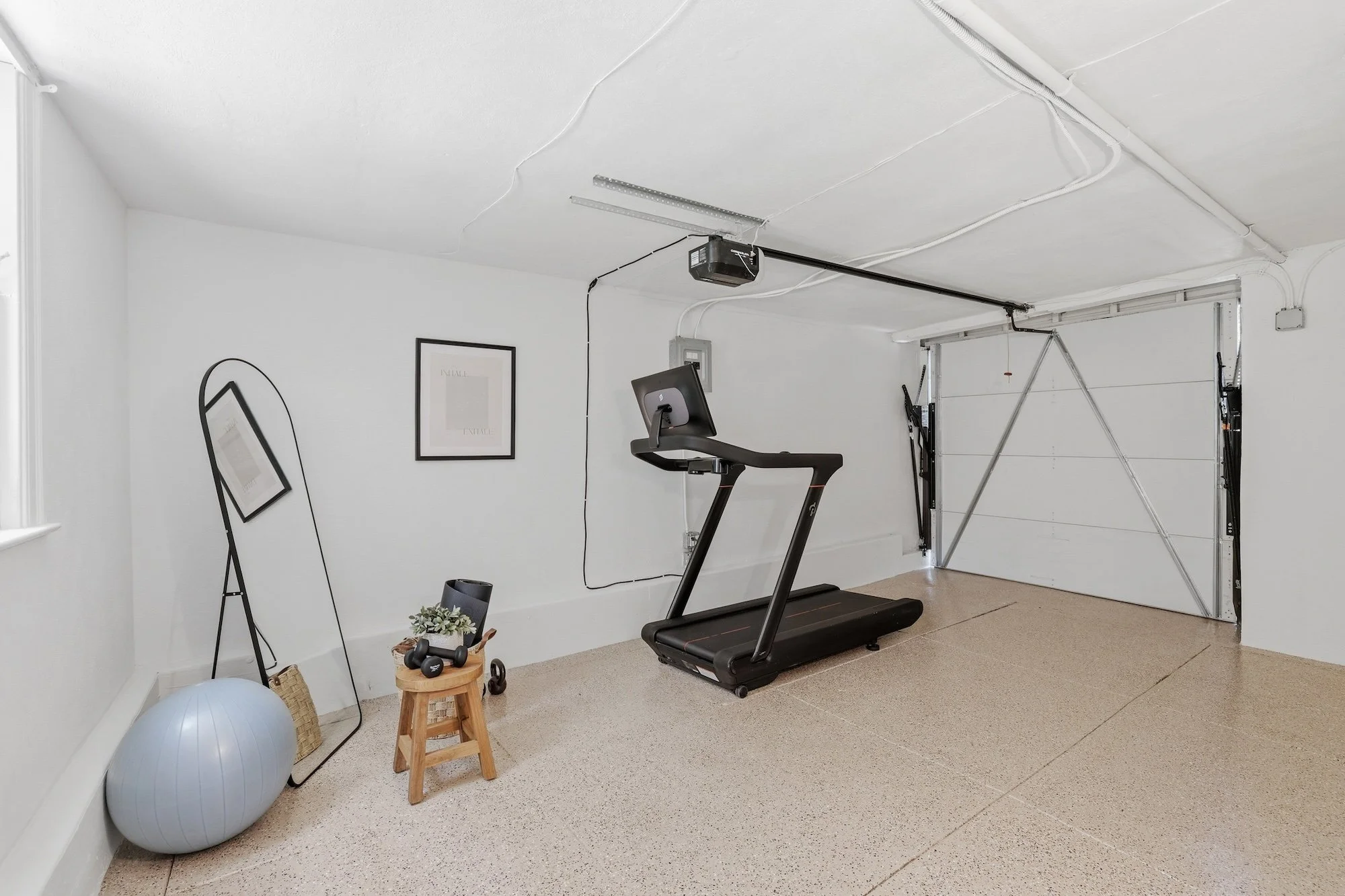 Home gym in a garage with a treadmill, exercise ball, small wooden stool, and wall art. The garage has a white door, shelves, and visible wiring.