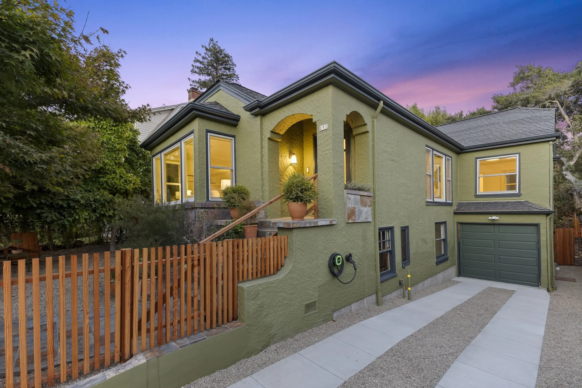 A two-story house painted in a light green color with dark gray trim. It has large windows with warm yellow interior lights on, and a garage with a dark green door. The front yard has a wooden fence, potted plants, and a paved walkway. There are tree