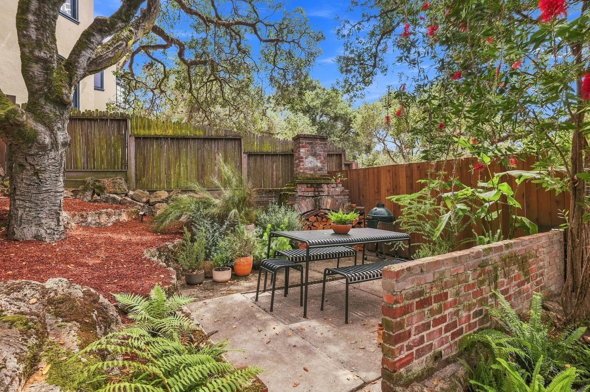 A backyard patio area with seating, plants, and a brick fence surrounded by tall trees and a blue sky.