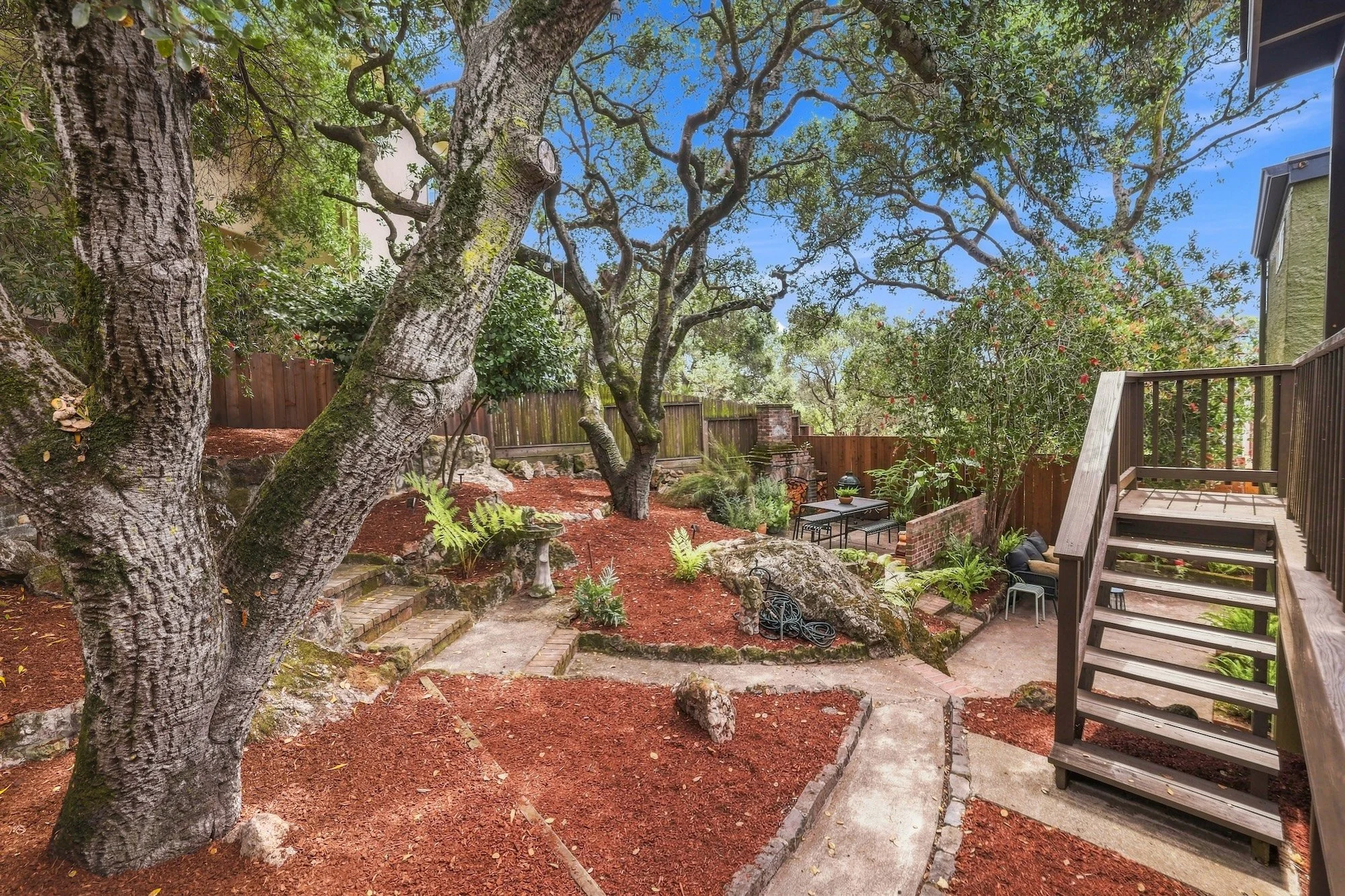Backyard garden with large trees, red mulch, stone pathway, garden furniture, and wooden fence.