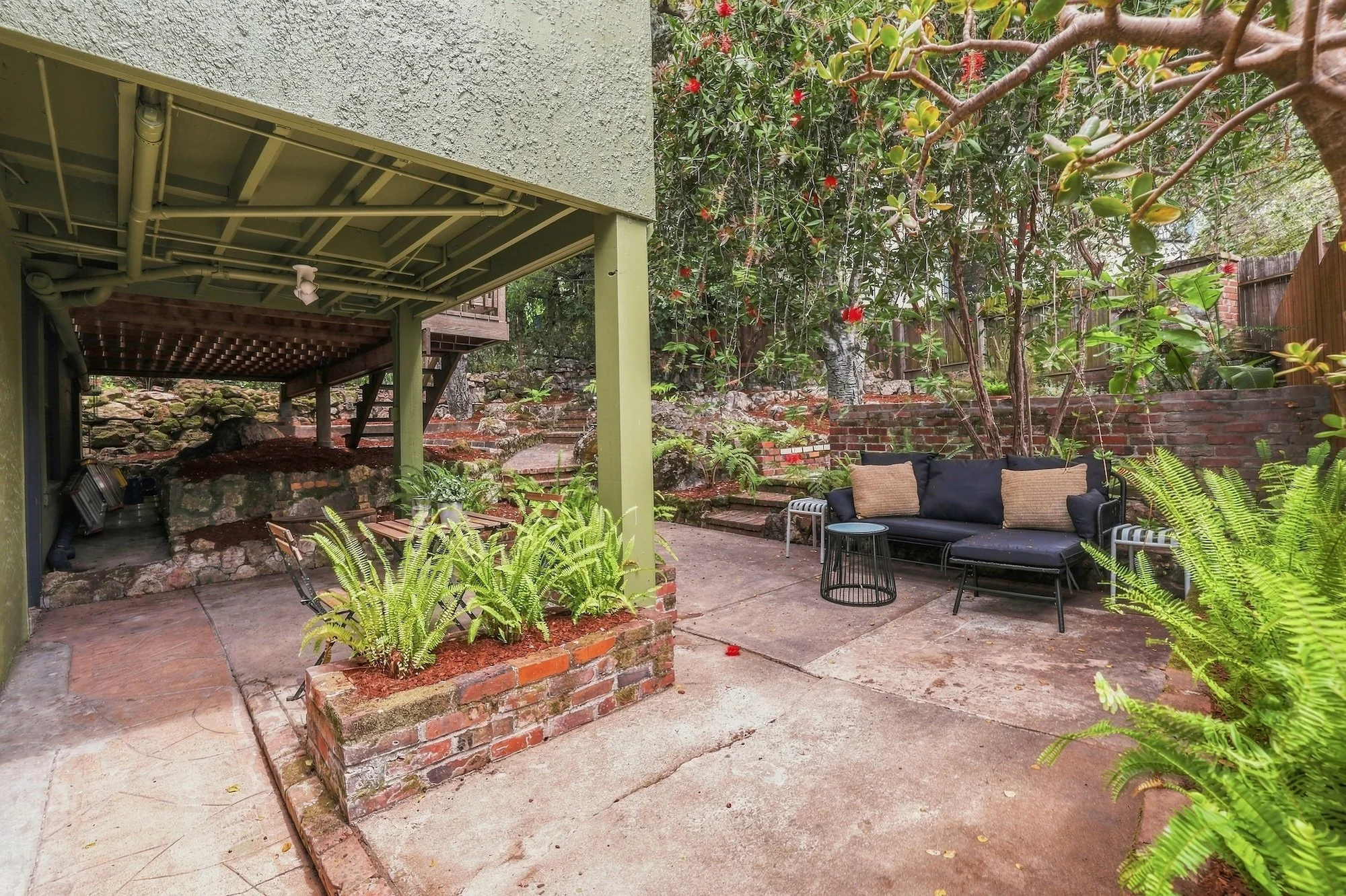 A backyard patio with a concrete surface, surrounded by green plants and trees. There is a modern black outdoor sofa with beige cushions, a small round black side table, and a larger round black table. The patio is next to a house with a green-painte