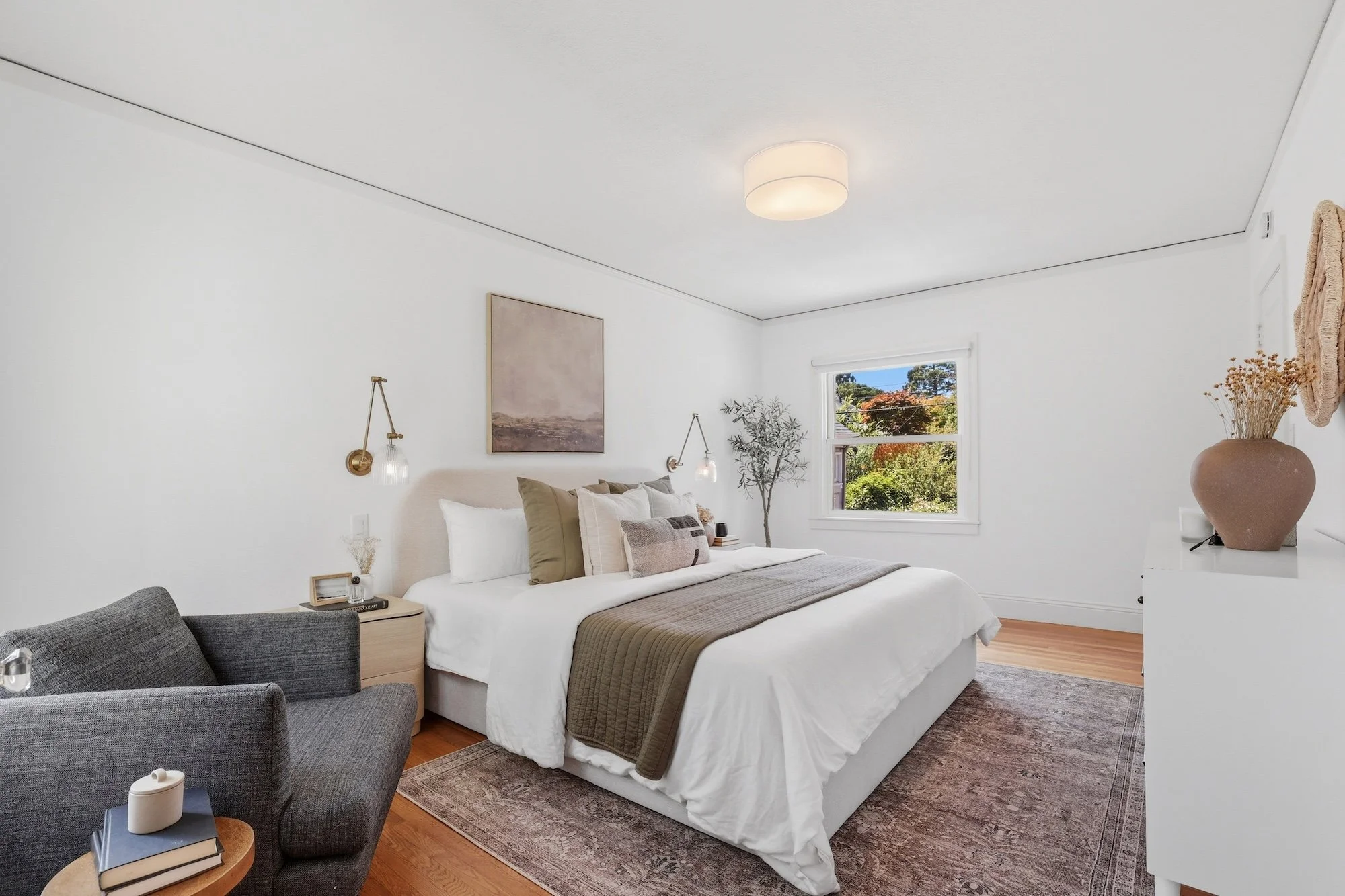 A bedroom with a white bed, beige pillows, and a brown blanket; a gray armchair nearby; a side table with books and candles; a large window showing trees outside; white walls with minimal decor; ceiling light fixture.
