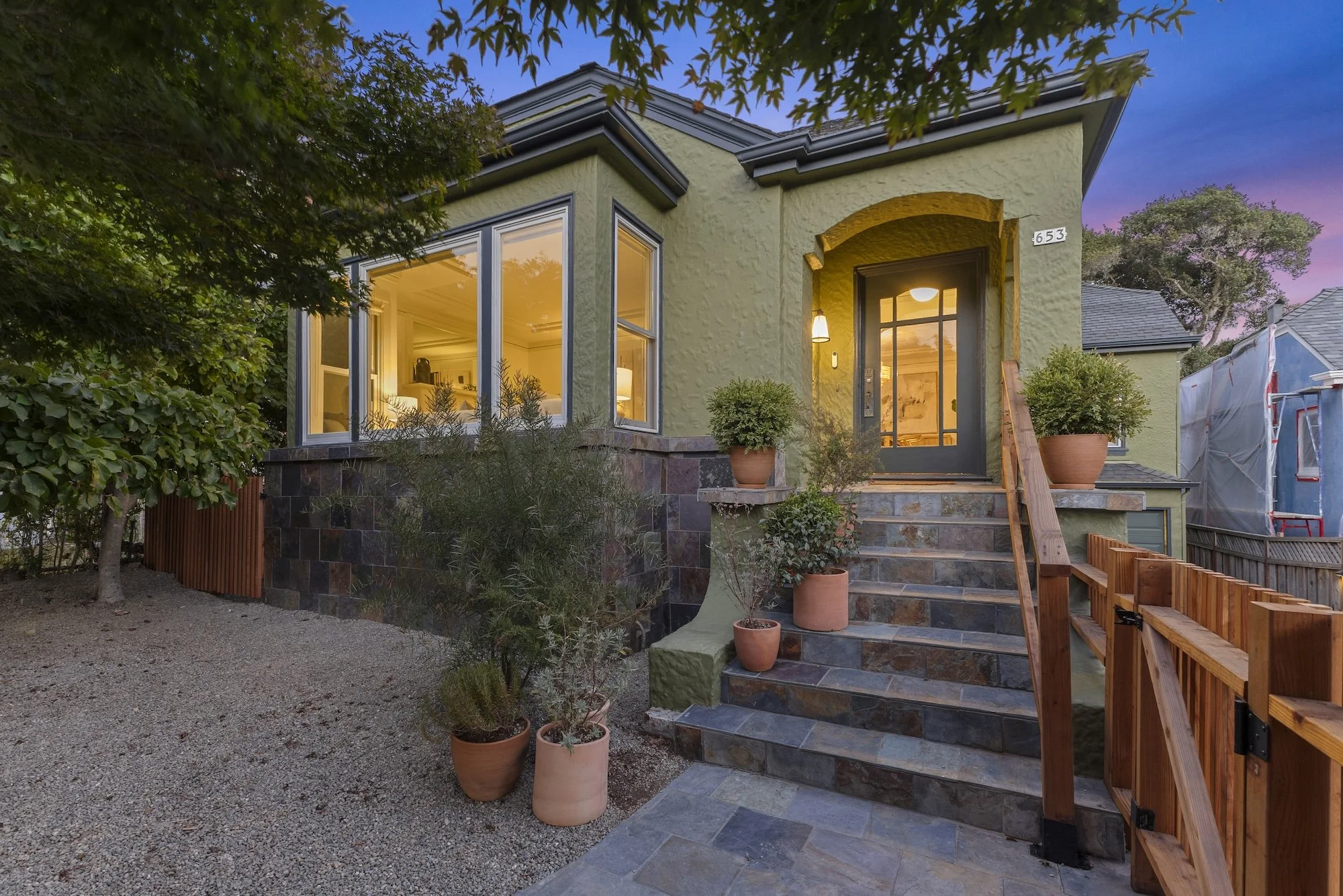 A two-story house with light green exterior walls, large front windows, and a dark gray door. The house has a stone staircase leading to the front door, decorated with potted plants on either side. The background shows a sunset sky with purple hues, 