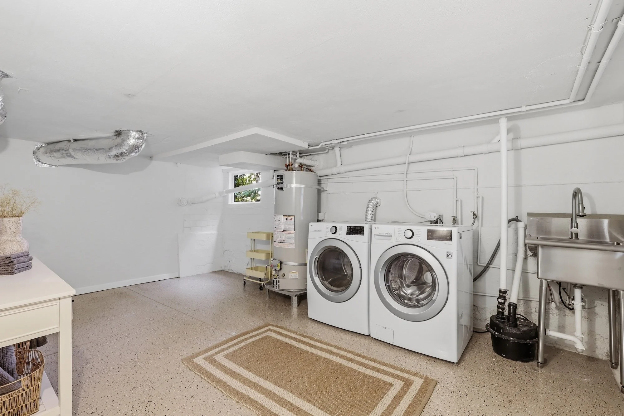 Laundry room with white walls and concrete floor, containing a front-loading washing machine and dryer, a water heater, a small window, and various pipes and ductwork on the ceiling.